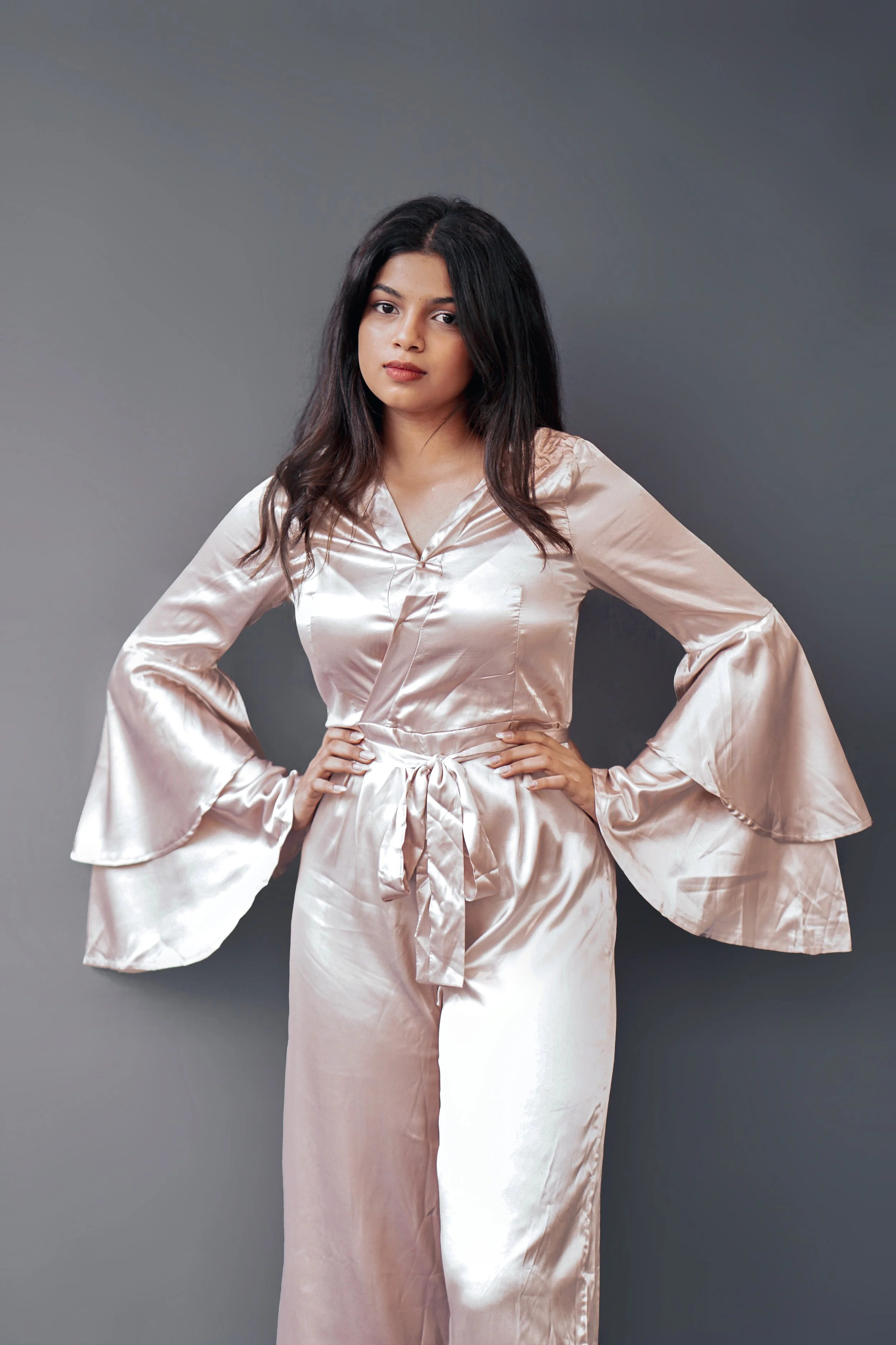 A woman with long dark hair wears a light pink silk satin pajama set with wide bell sleeves and stands against a plain gray background.