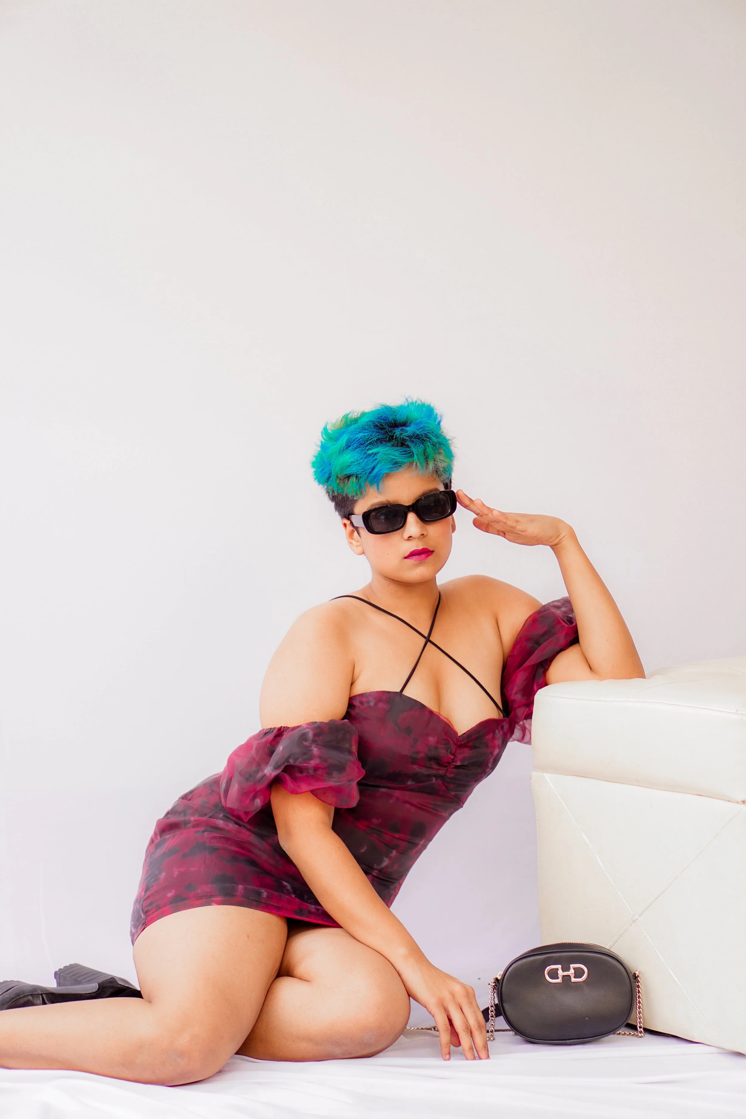 A woman with short, colorful hair in shades of blue and green, wearing sunglasses, a dark red floral dress, and black shoes, kneeling on a bed next to a white headboard and a black purse.