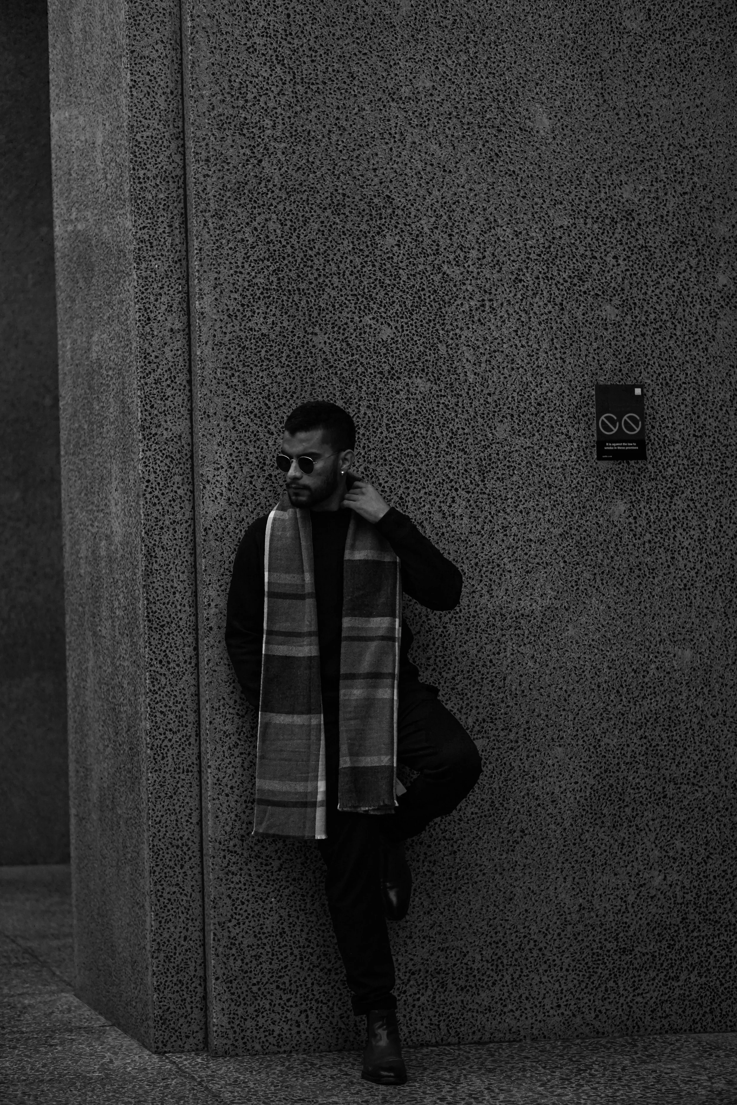 A man with sunglasses, a scarf, and dark clothing leaning against a textured wall, standing on one leg with the other foot raised and bent at the knee.