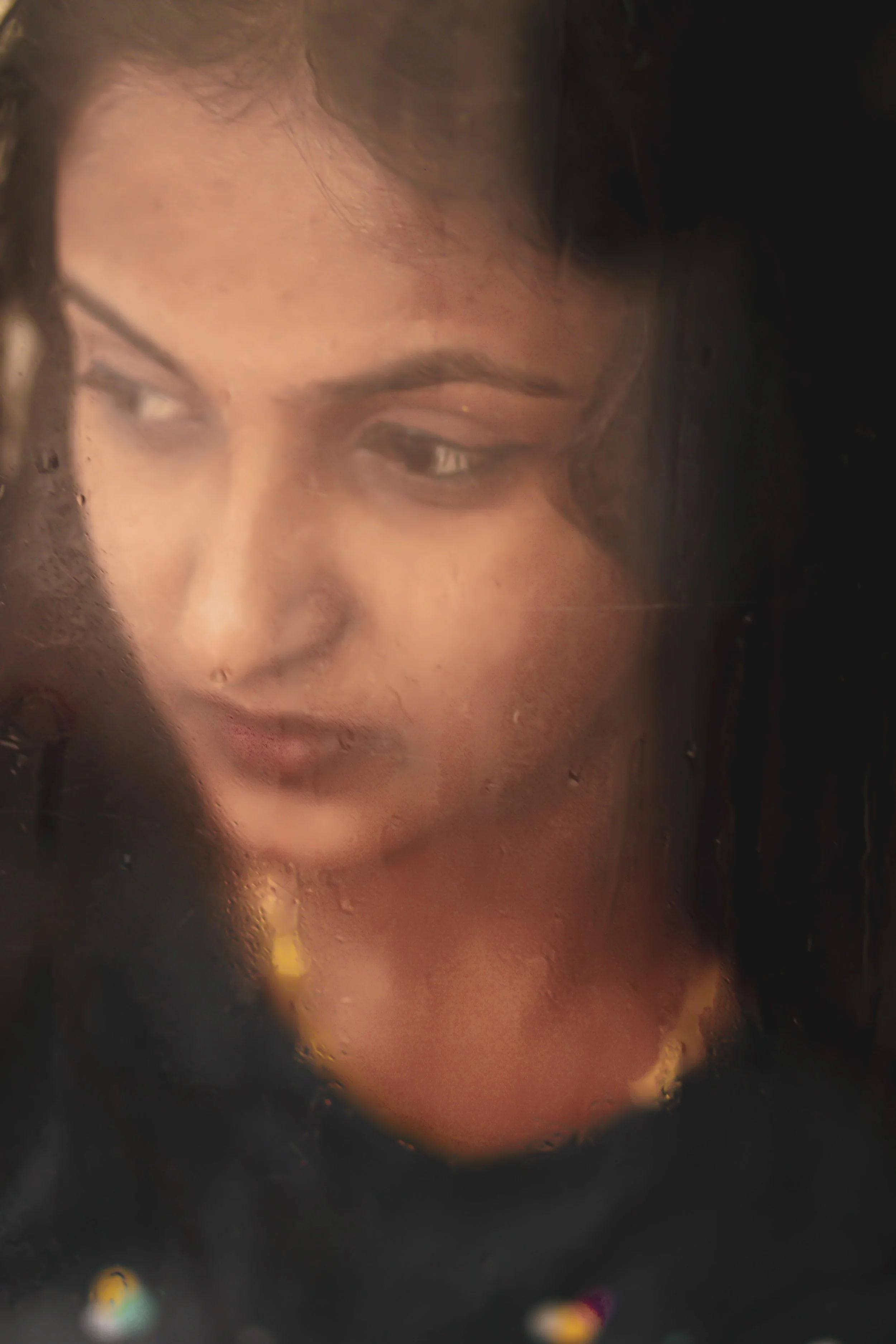 A woman's face seen through a foggy or dirty glass surface, with water droplets visible on the glass, giving a blurred and obscured view.