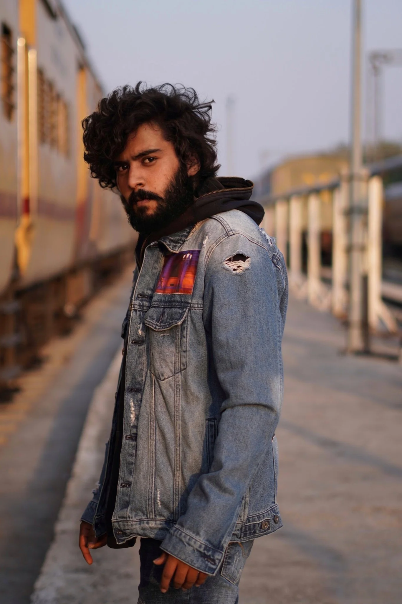 A young man with dark curly hair and a beard standing on a train platform, wearing a distressed denim jacket layered over a hoodie.