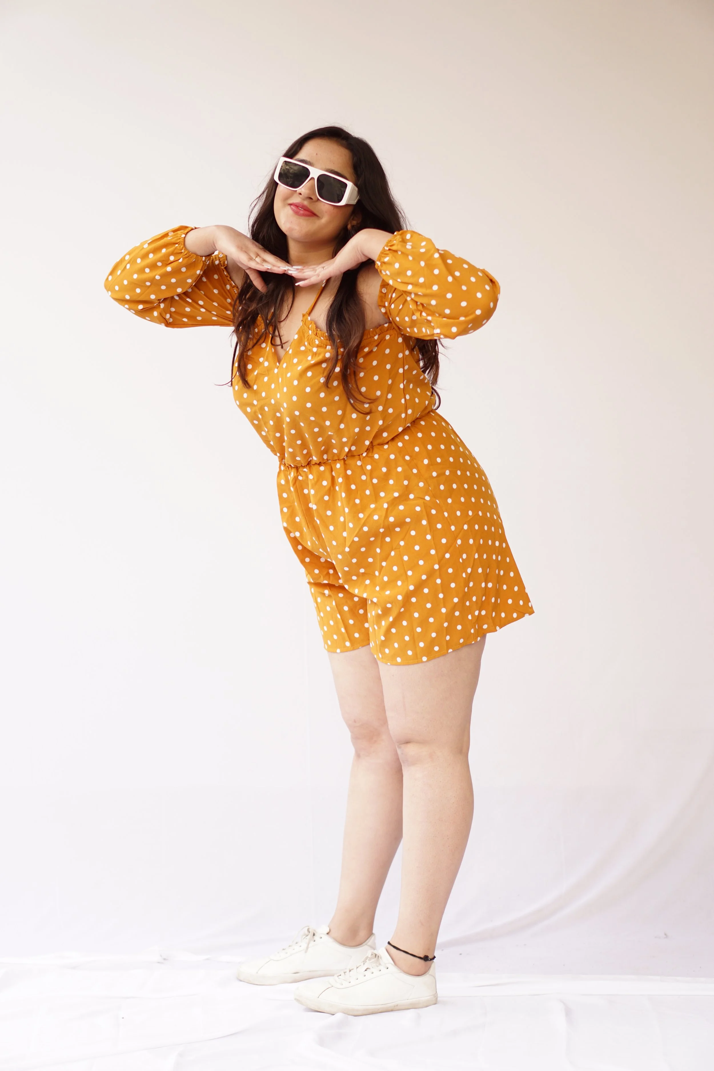 A woman in a mustard yellow polka dot romper, wearing white sneakers and oversized white sunglasses, posing with a playful expression against a plain white background.