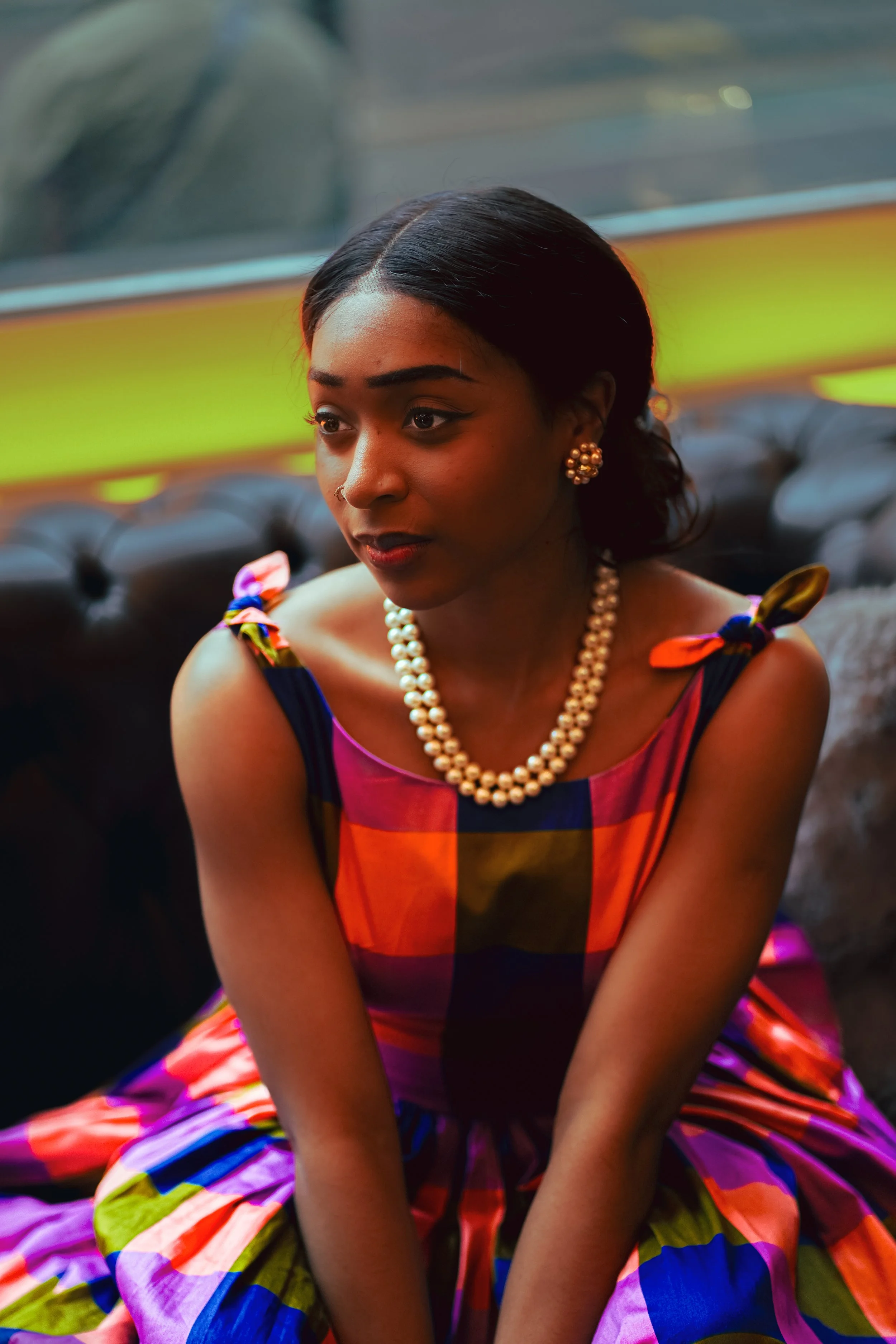 A woman wearing a colorful checkered dress with tied shoulder straps, pearl necklaces, and earrings, sitting on a black leather sofa inside a dimly lit room with neon lighting visible in the background.