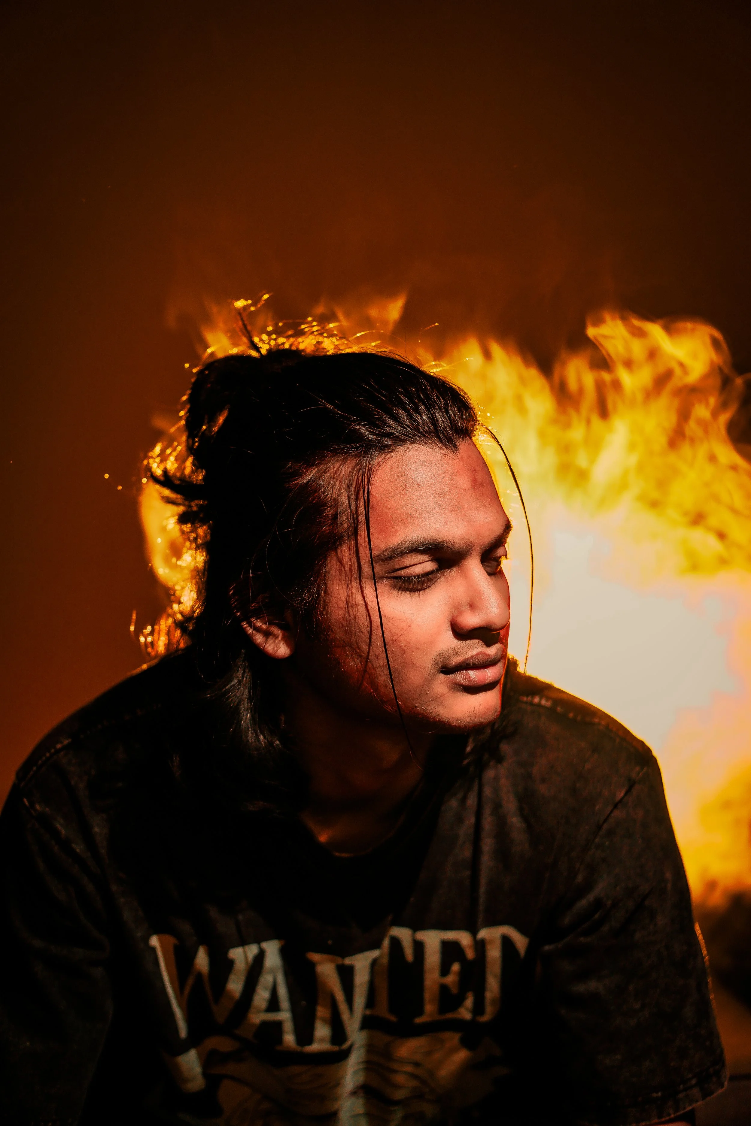 A young man with long dark hair, closed eyes, and wearing a black shirt with 'WANTED' written on it, poses in front of a fire with orange flames and dark background.
