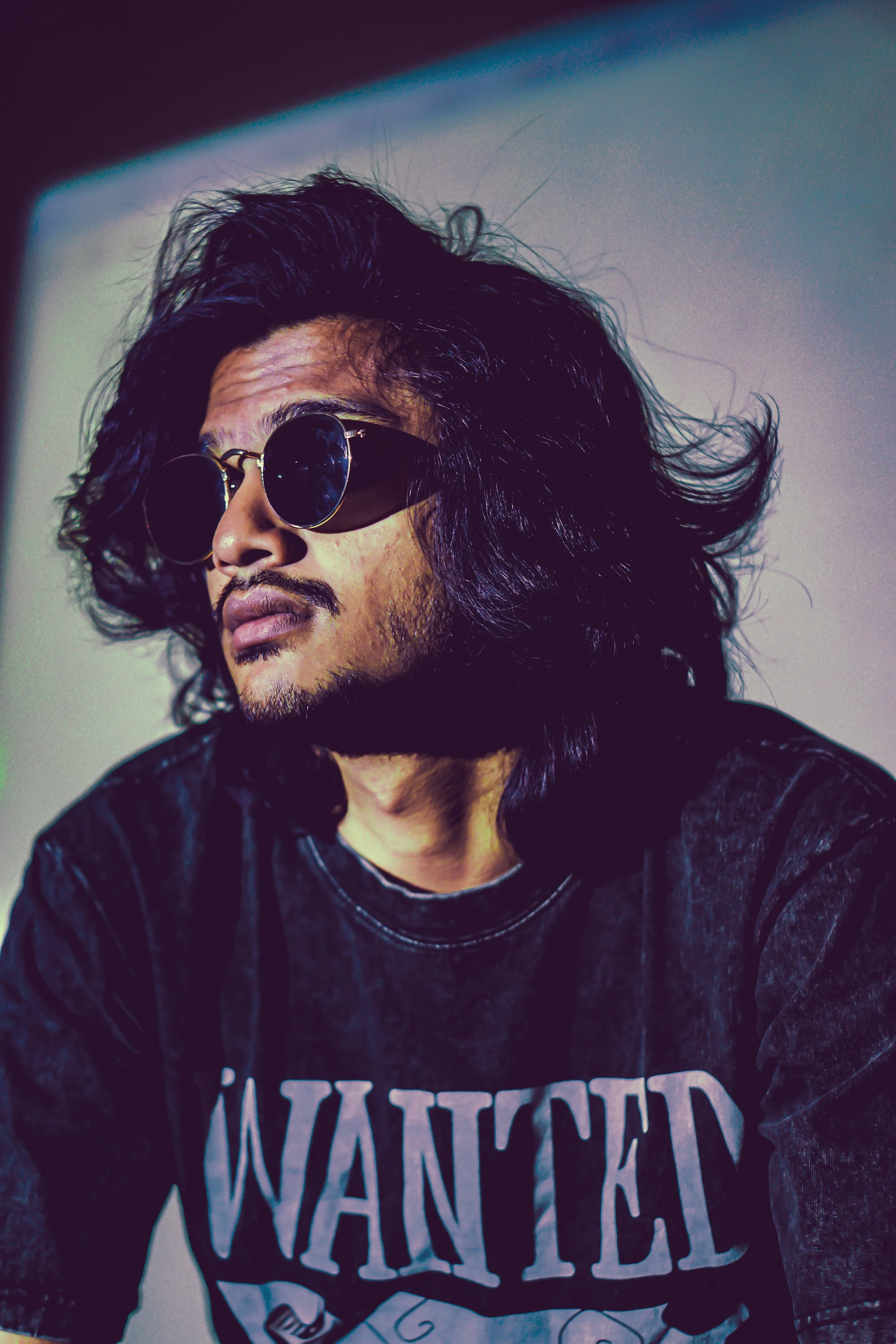 A man with long dark hair, wearing round sunglasses and a black shirt with the word 'WANTED' printed on it.