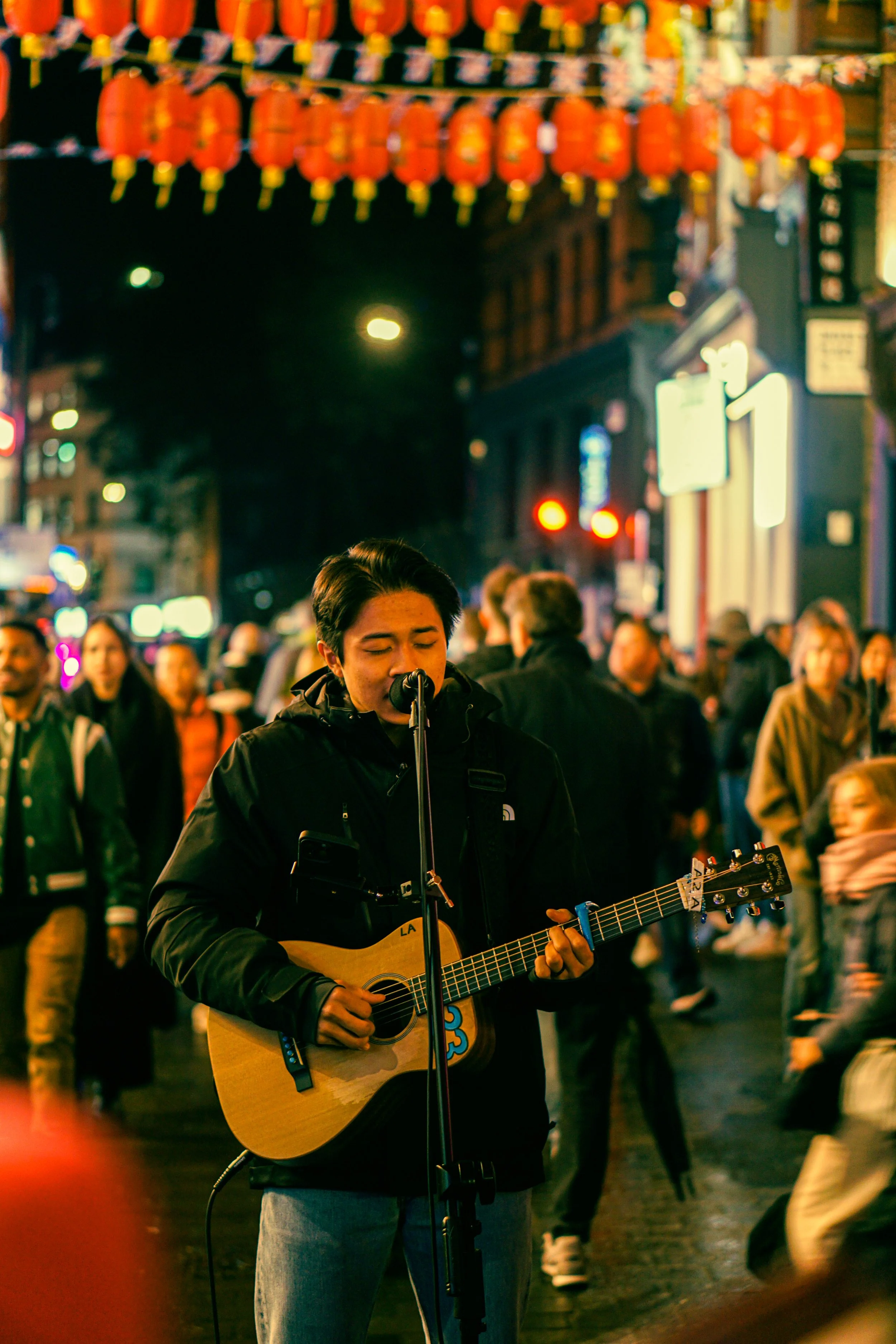 A young man with dark hair, wearing a black jacket, playing an acoustic guitar and singing into a microphone on a busy street at night. The street is decorated with red lanterns hanging above, with many people walking in the background.