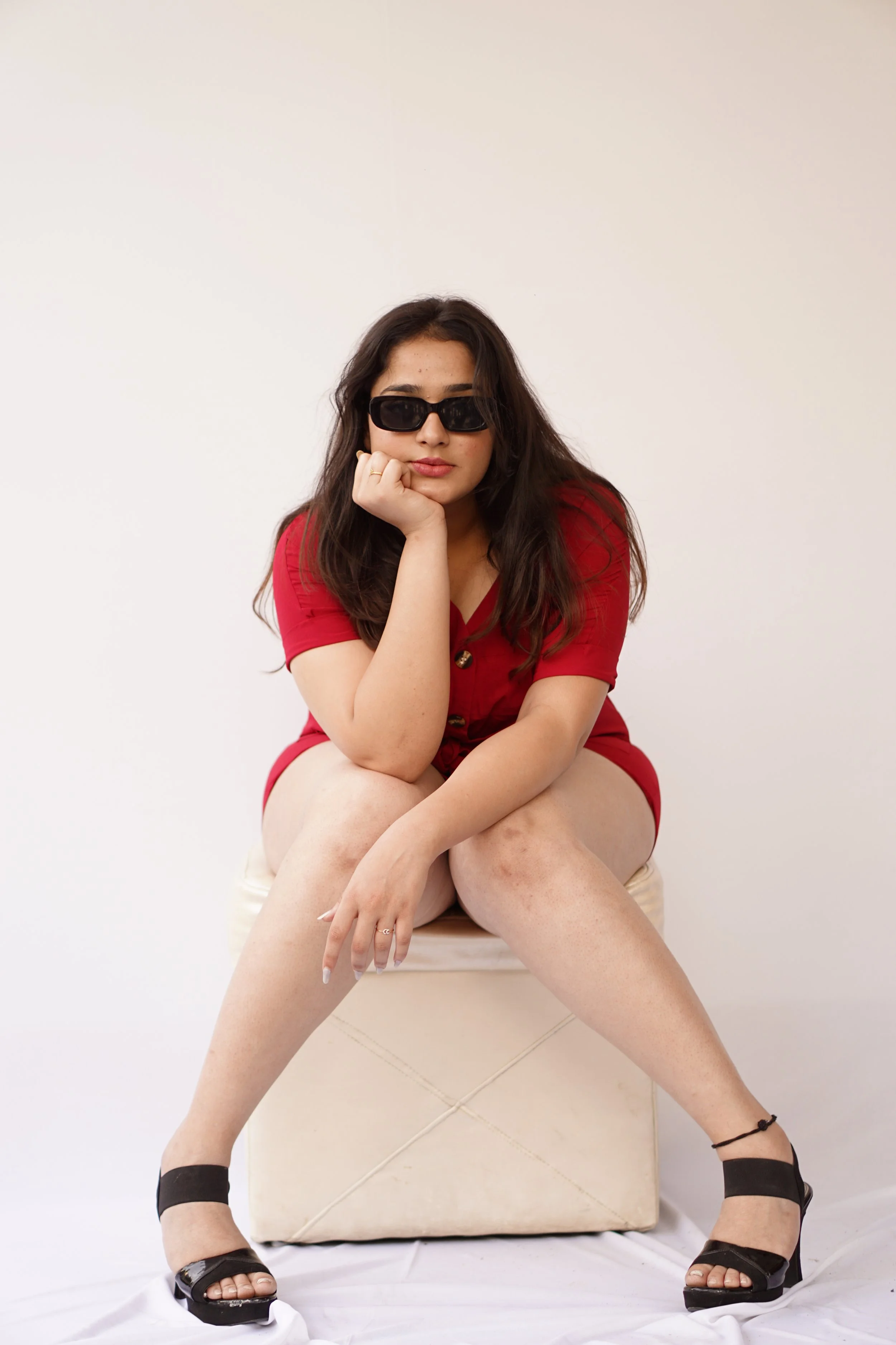 Young woman wearing black sunglasses and a red dress, sitting with her legs apart on a cushioned stool against a plain white background.