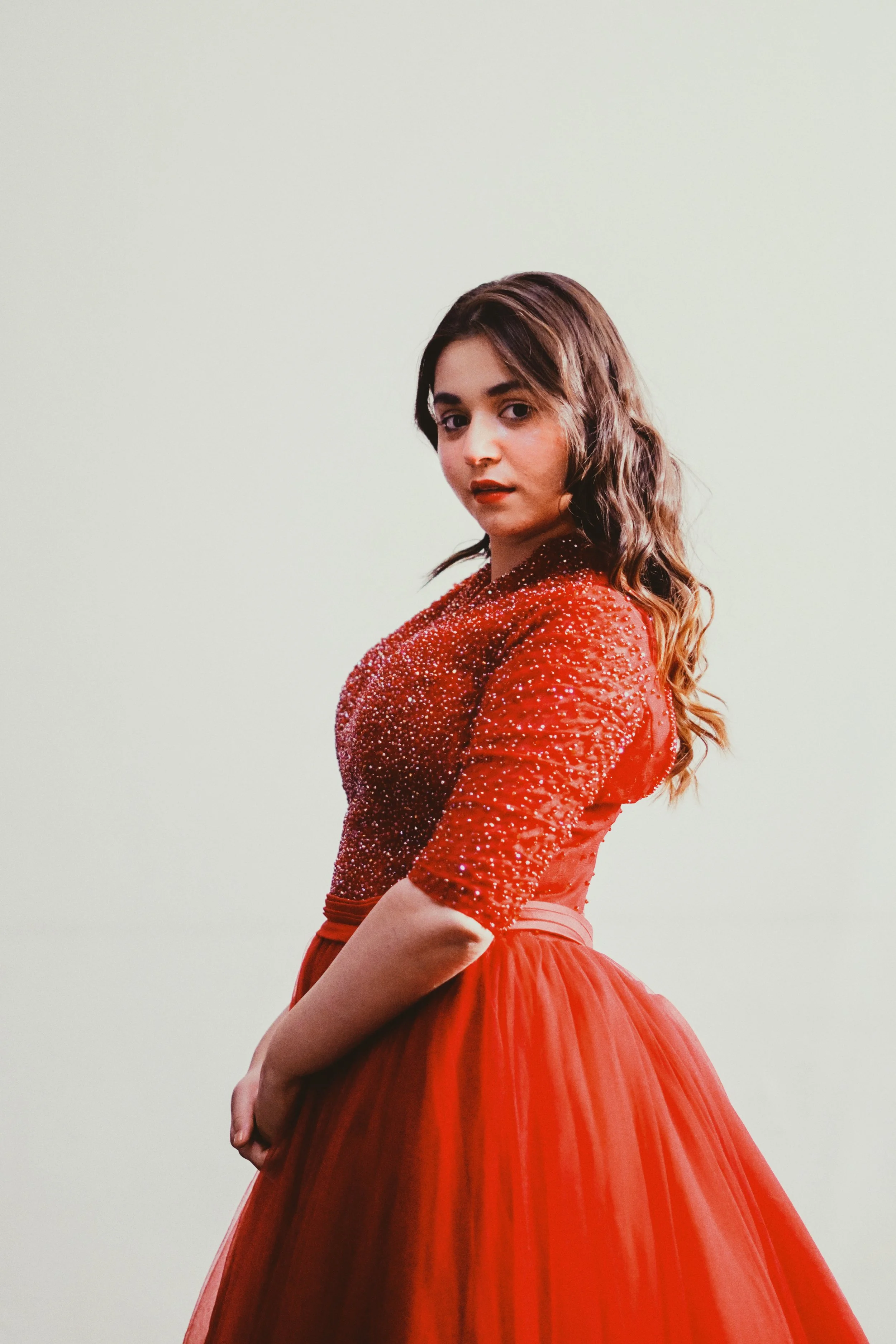 A woman with wavy brown hair wearing a red, sparkly dress with three-quarter sleeves and a full skirt, standing against a plain light background.