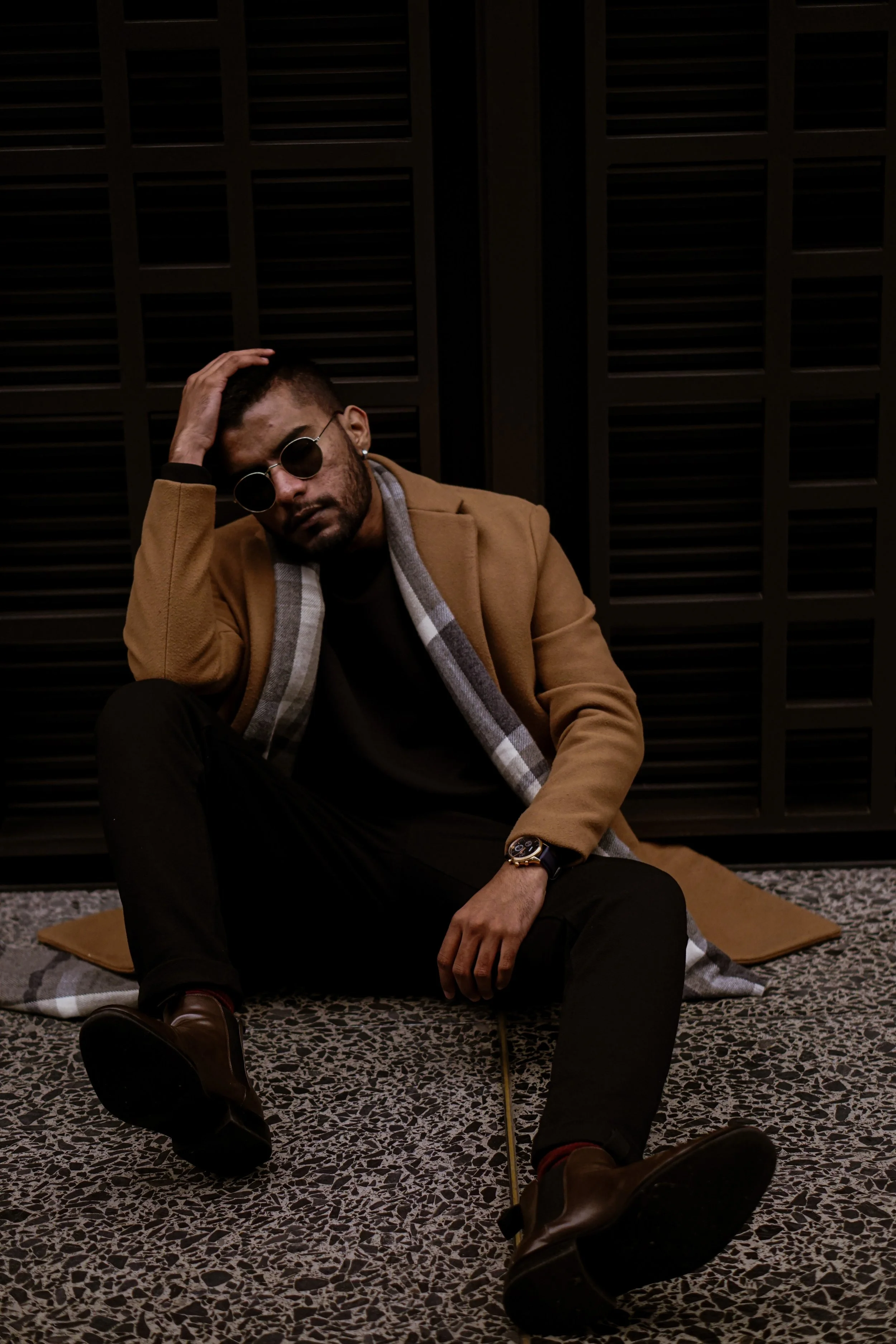 A man sitting on the floor in front of a dark background, wearing sunglasses, a brown coat, a scarf, black pants, and brown shoes, with one hand on his head and the other resting on his knee.