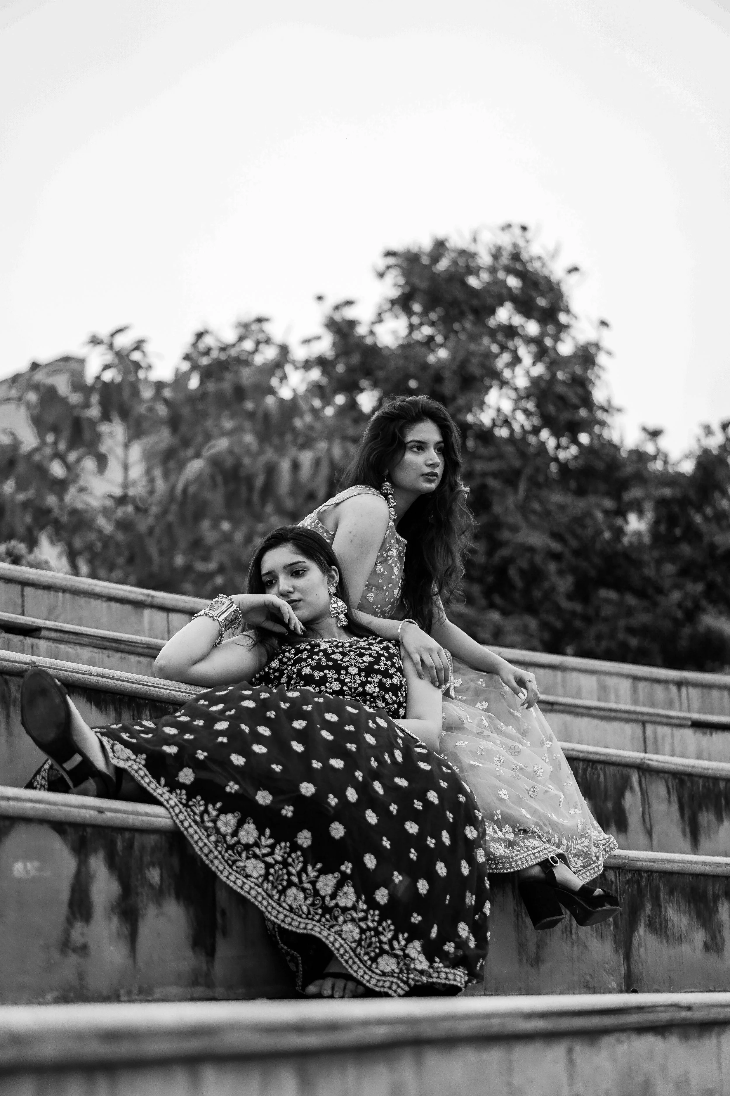 Two women dressed in traditional Indian clothing are sitting on outdoor stairs, one resting her head on the other's lap; they appear contemplative.