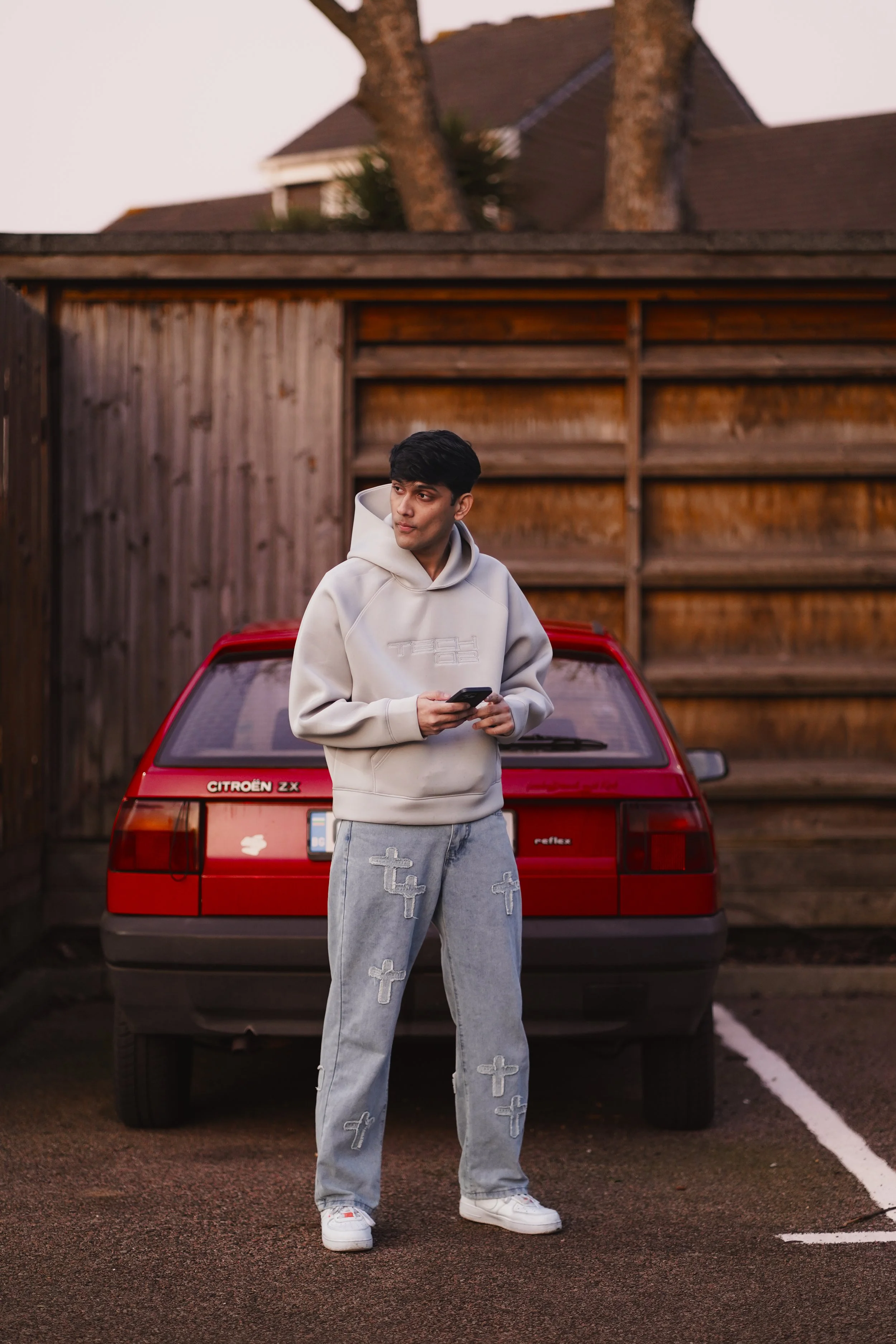 A young man in light gray hoodie and jeans with cross patches, holding a phone, standing in front of a red Citroën ZX car in a parking lot with a wooden fence and house in the background.