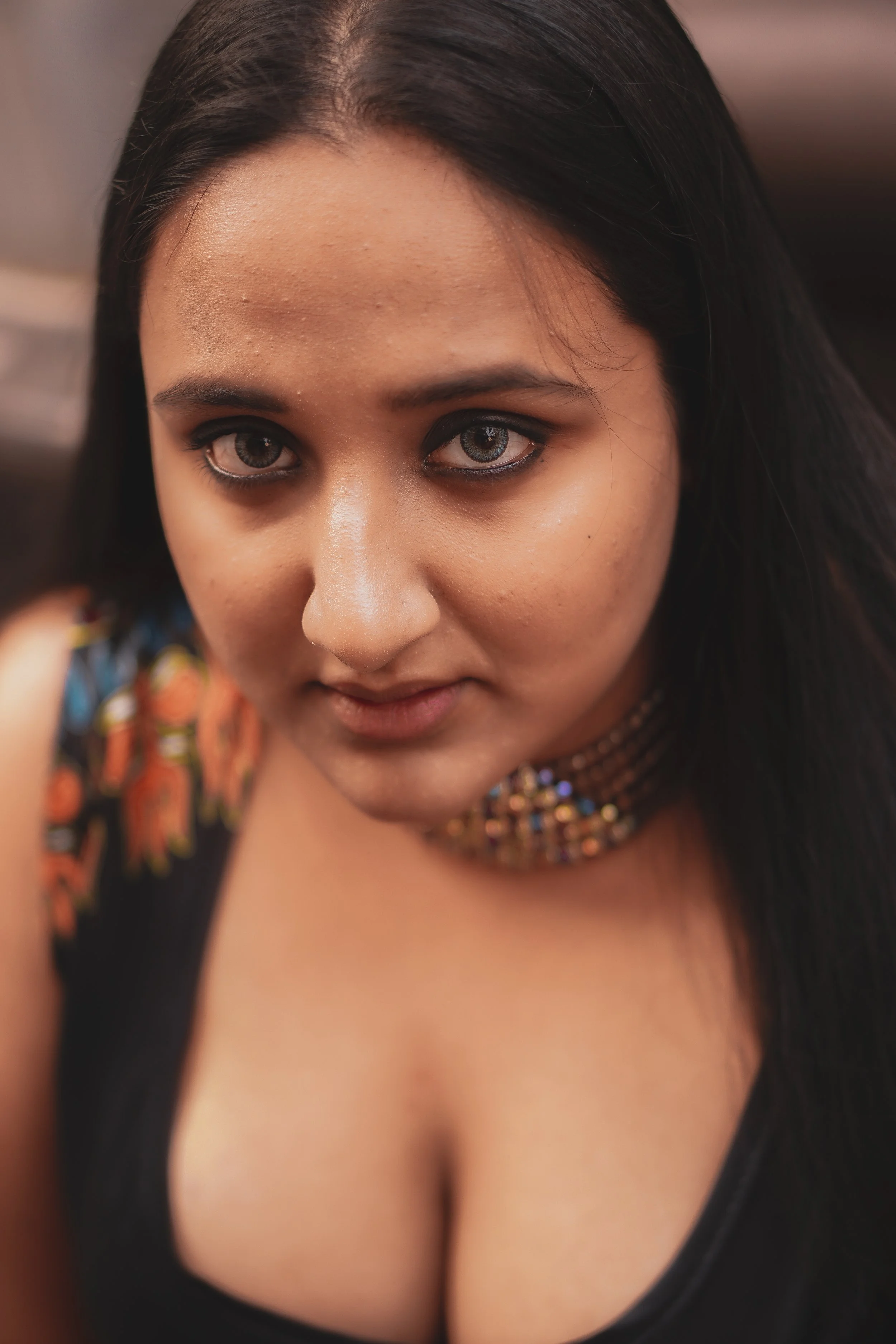 A young woman with long dark hair, wearing makeup with light blue eyes and dark eye makeup, a colorful beaded necklace, and a black top with a floral pattern on the shoulder.