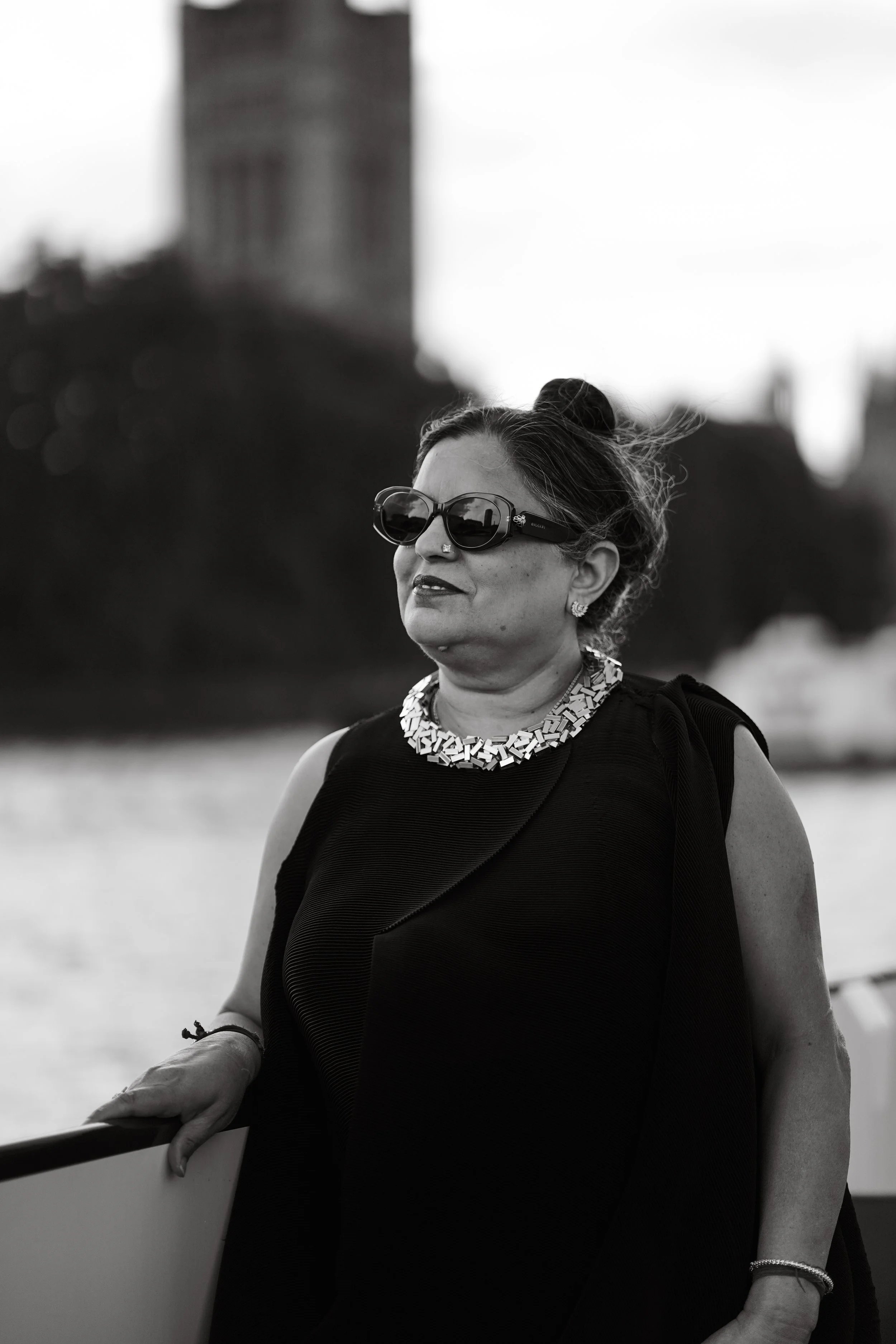 A woman in black sleeveless top, wearing sunglasses and jewelry, standing outdoors against a blurred backdrop of water and distant buildings.