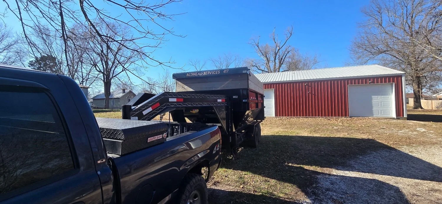 Black pickup truck towing a dump trailer parked on a grassy area in front of a red and white metal garage