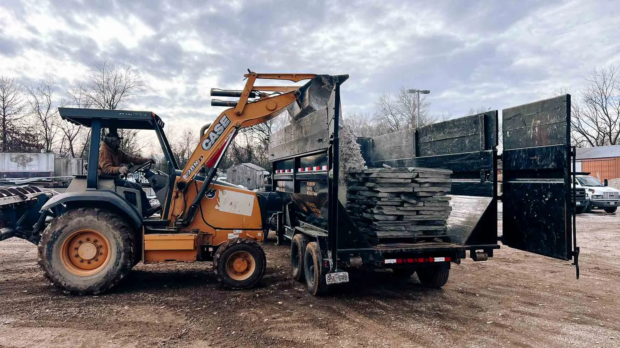 A front loader with a CASE logo is unloading slate tiles from a black trailer in a gravel yard. The sky is partly cloudy with leafless trees in the background.