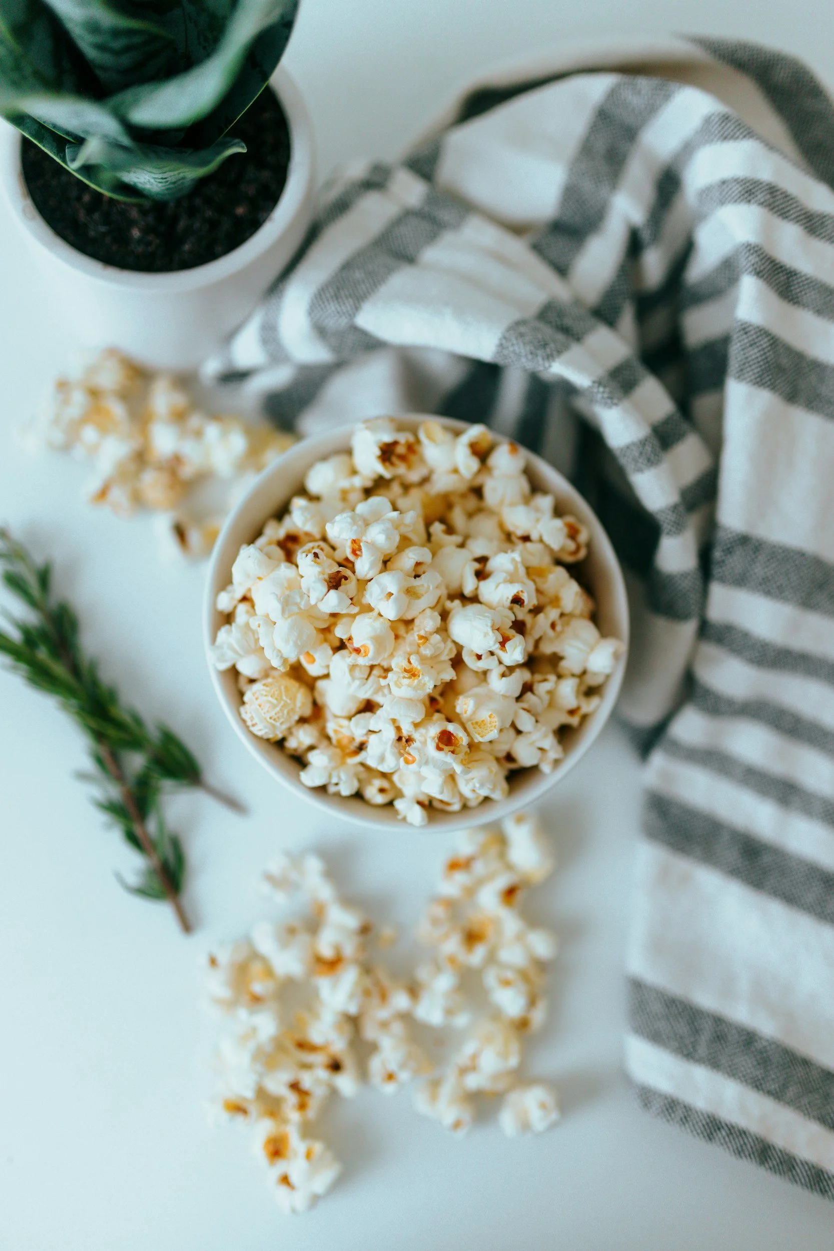 A bowl of popcorn on a white surface with a sprig of rosemary, a potted plant, and a striped cloth nearby.