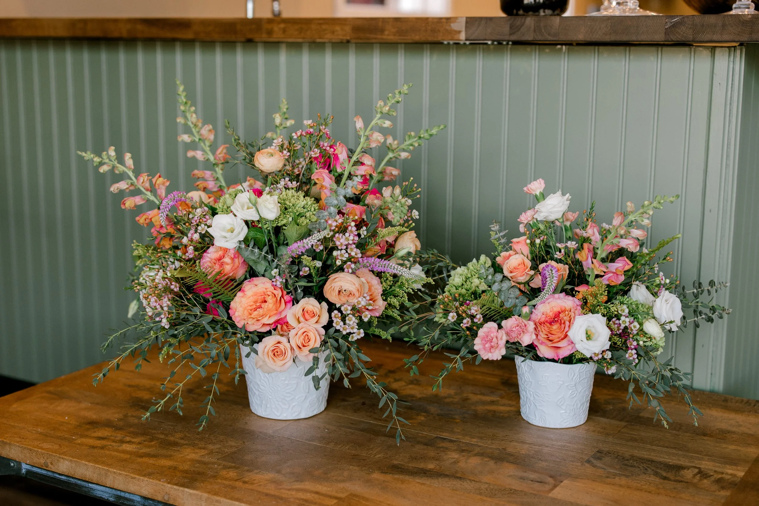Two white floral arrangements in white textured pots on a wooden table with a green paneled wall in the background.