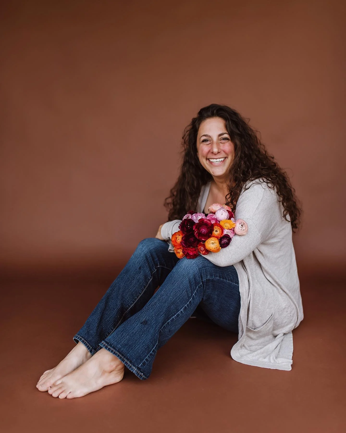 Woman with curly hair sitting on the floor, holding a bouquet of pink, orange, red, and burgundy flowers, smiling, wearing a gray cardigan and blue jeans, against a brown background.