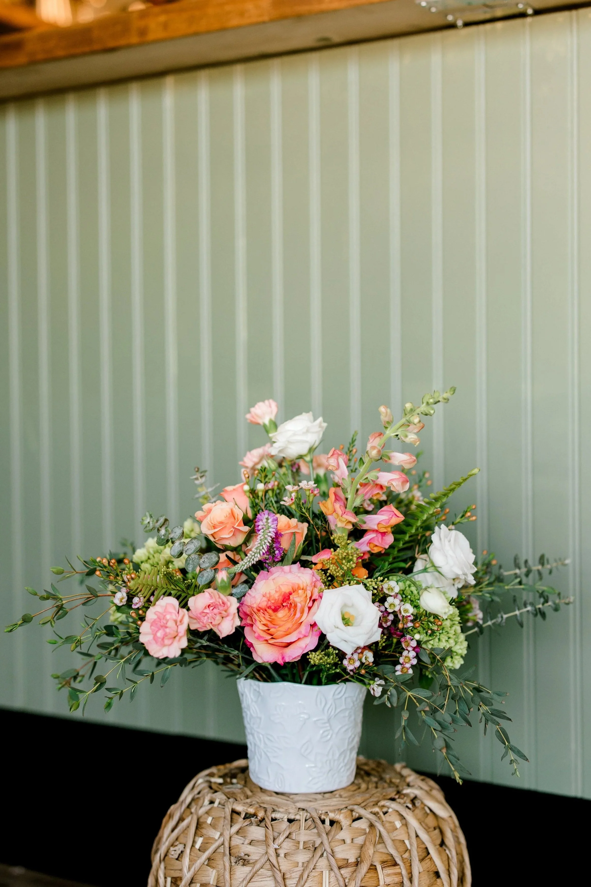 A colorful bouquet of flowers in a textured white vase on a woven wicker table against a pale green beadboard wall.