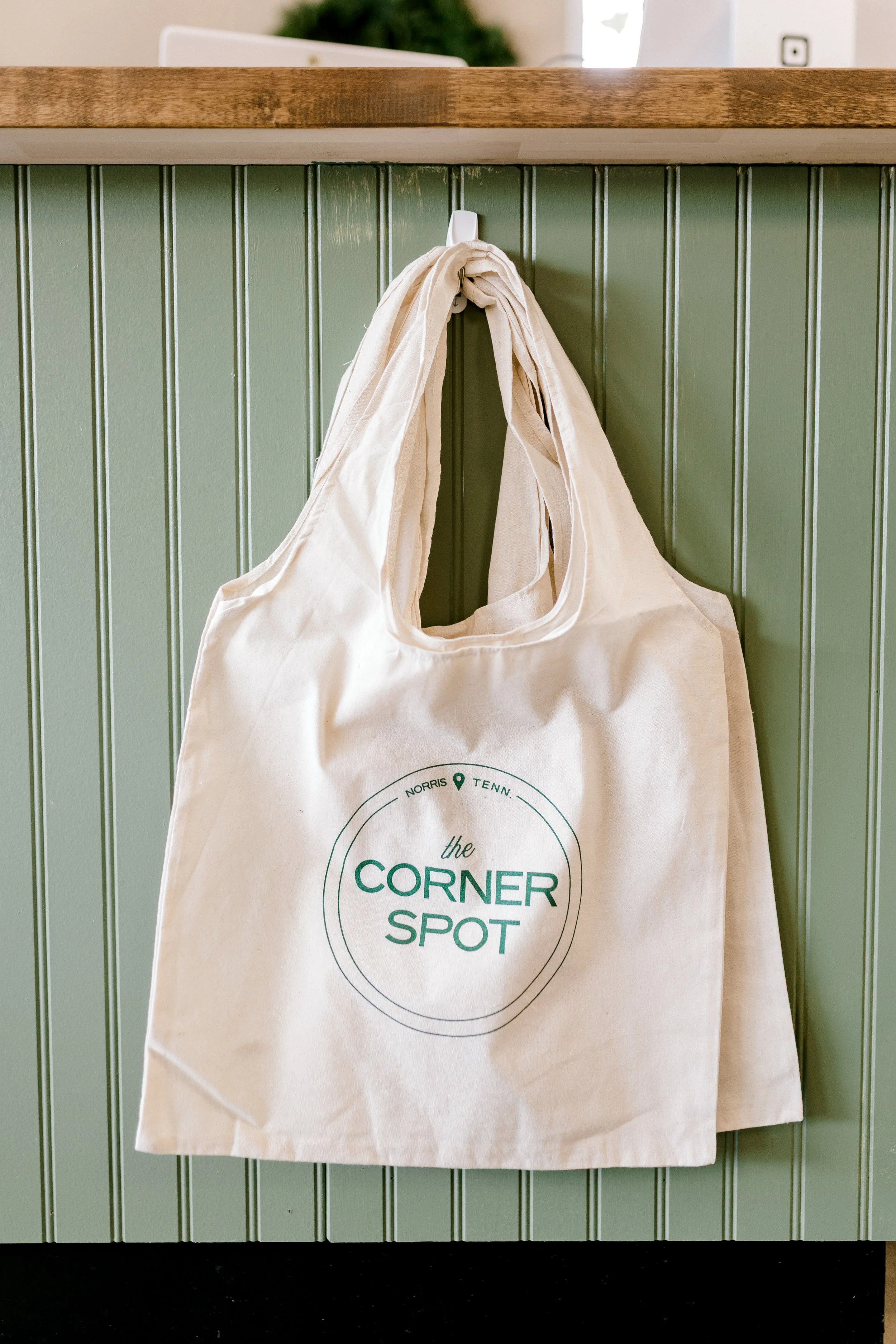 Reusable fabric bag hanging on a hook, with 'the CORNER SPOT' logo printed on it, in front of a green paneled wall.