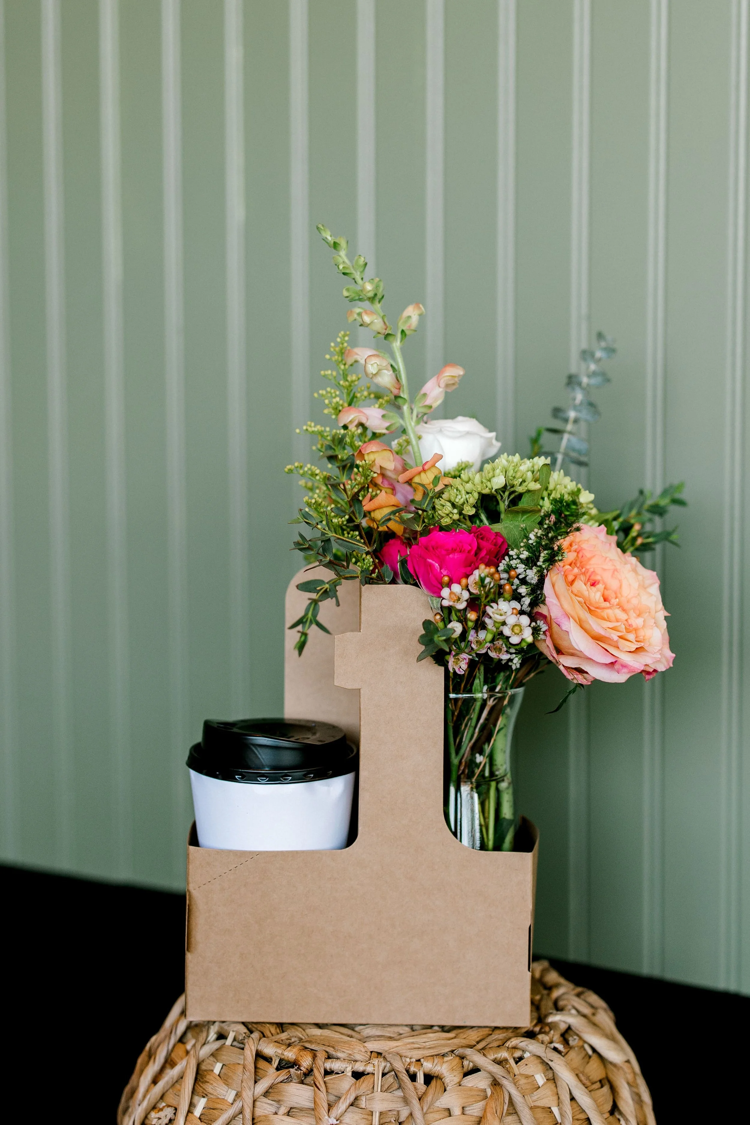 A bouquet of various flowers including pink and white roses, pink carnations, and greenery in a glass vase on a wicker table, with a coffee cup in a cardboard container beside it, against a green paneled wall.