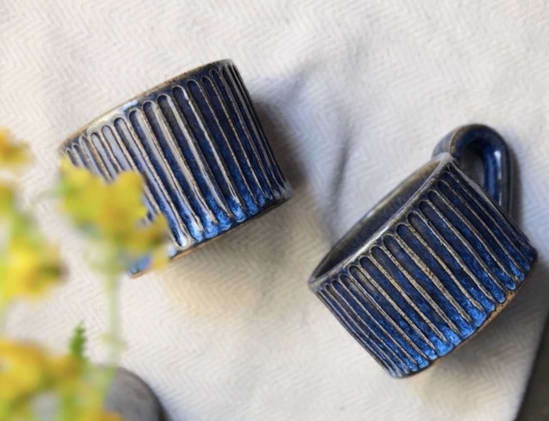 Two dark blue ceramic mugs with vertical ridges, placed on a light-colored fabric surface, with a blurred yellow flower in the foreground.