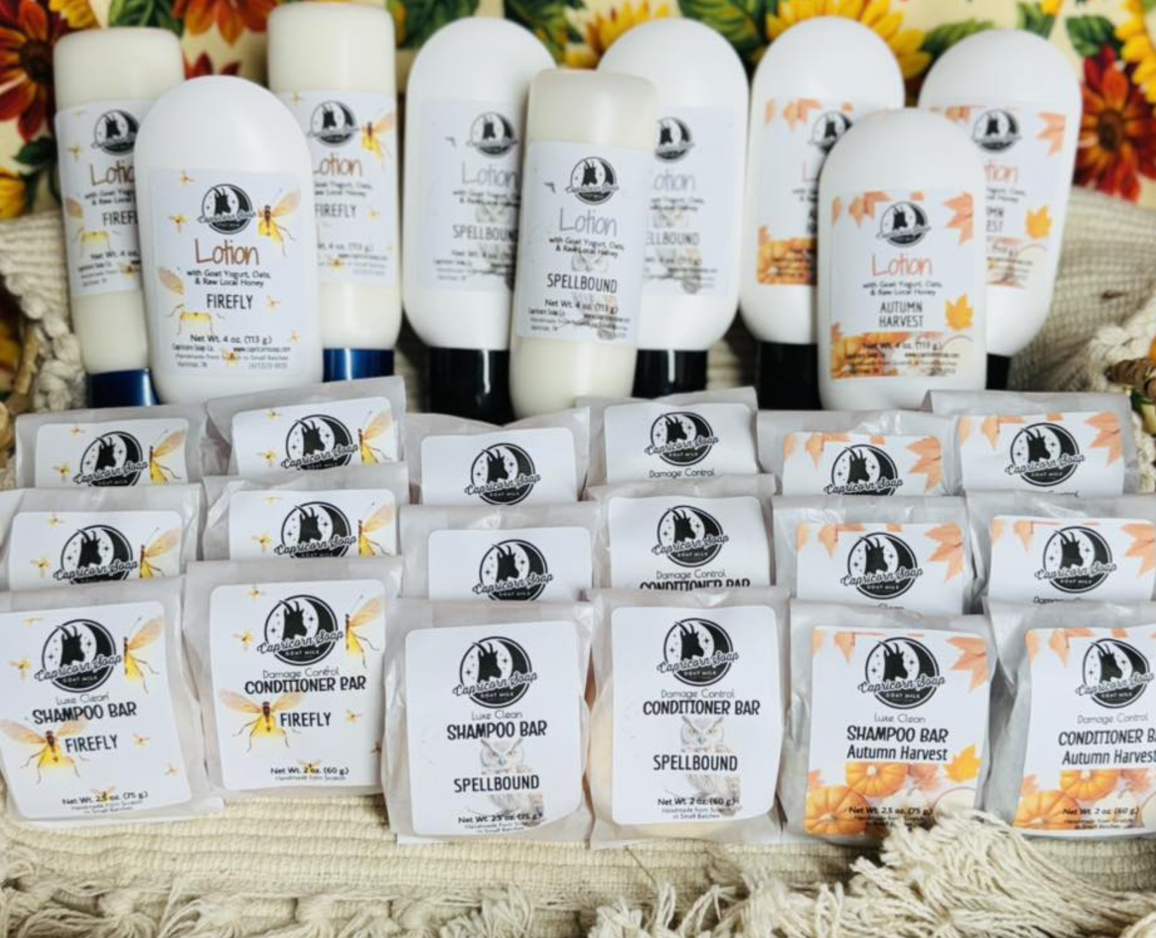 A display of handmade soap and lotion bottles with fall-themed labels, featuring scents like Firefly, Spellbound, and Autumn Harvest, arranged on a woven fabric surface with decorative flowers and foliage in the background.