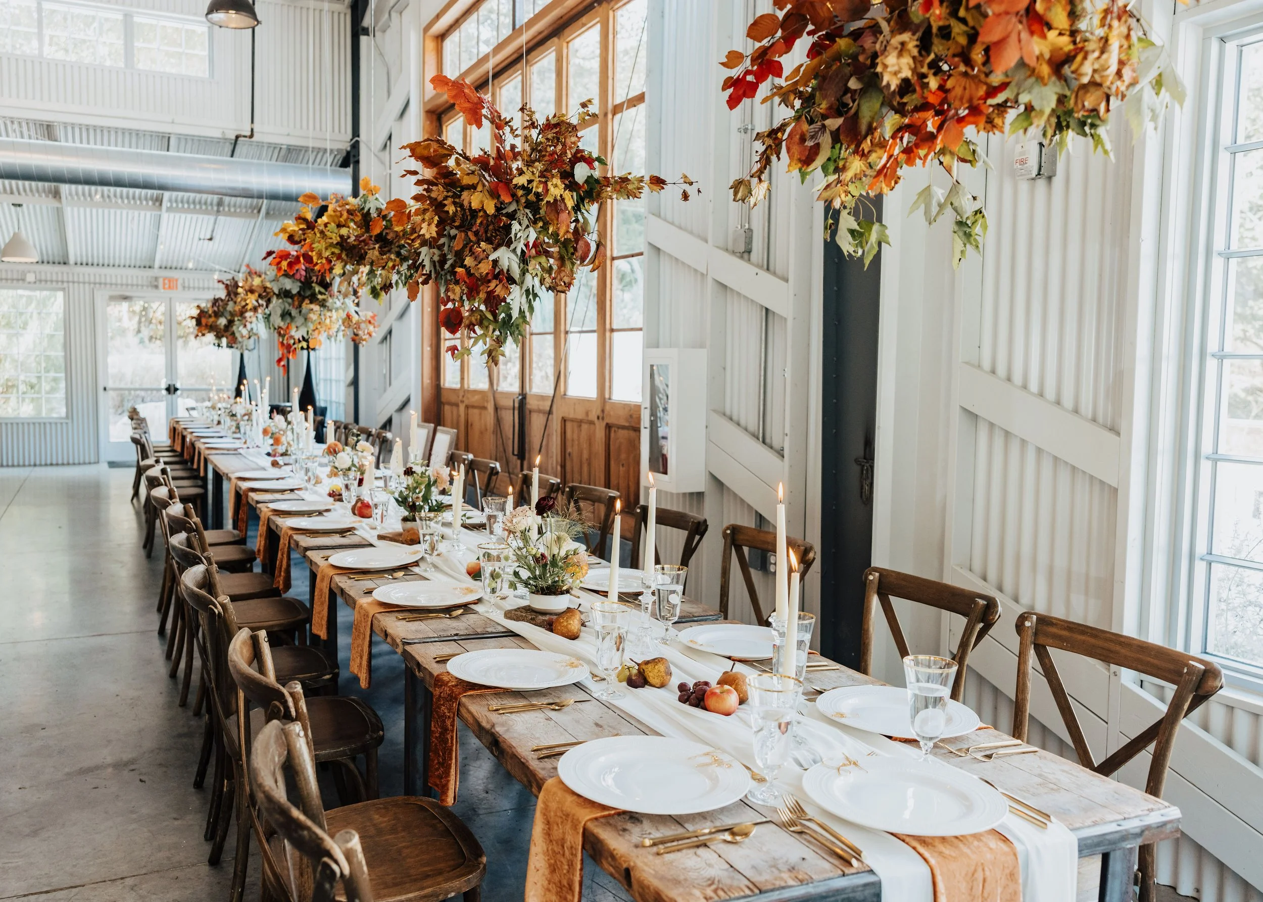 Long rustic wooden dining table set for a meal with white plates, gold utensils, wine glasses, and floral centerpieces. Overhead, autumn-themed foliage garlands are hung. The room has large windows, white wood-paneled walls, and a high metal ceiling.
