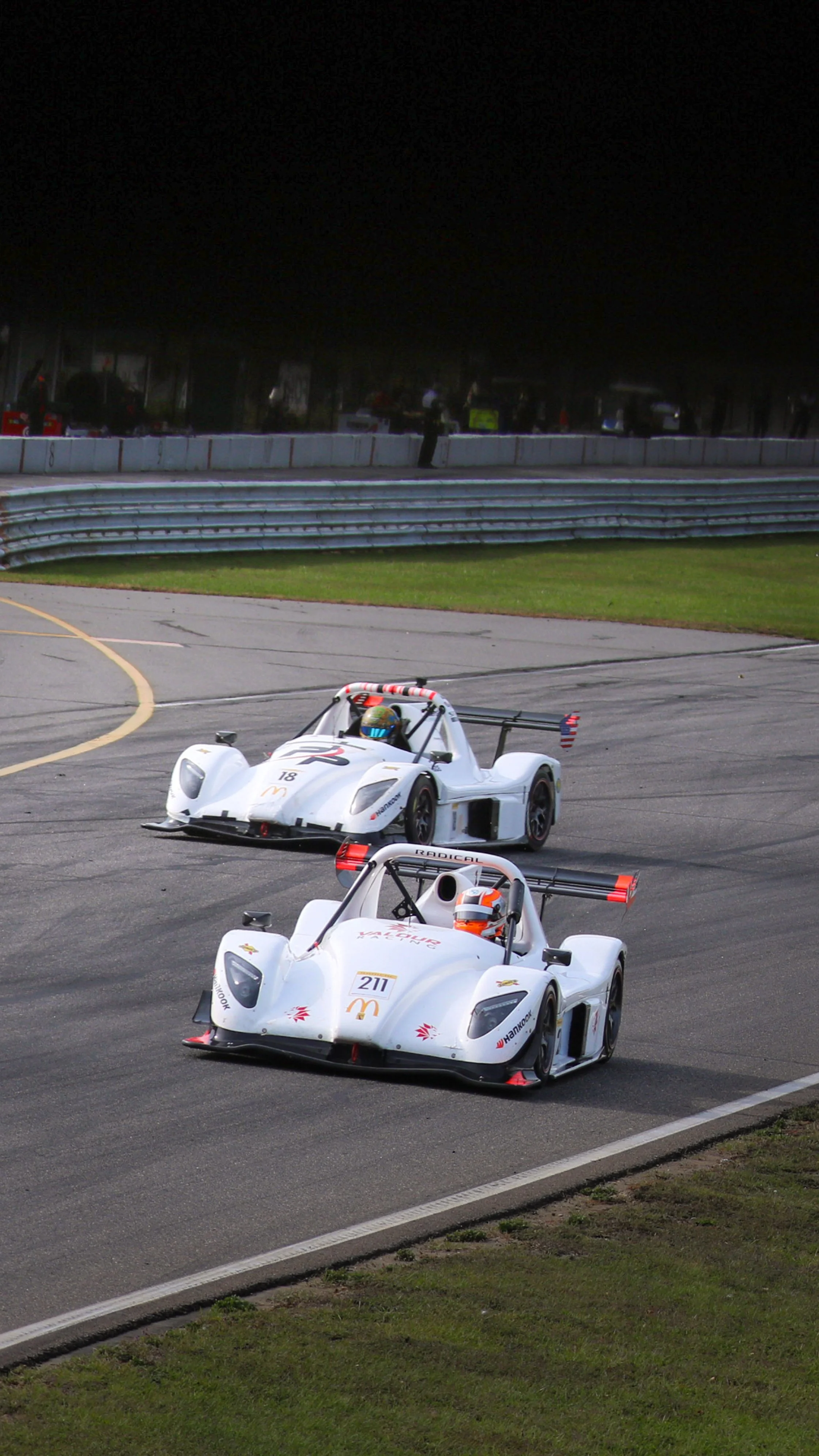 Two race cars, white with racing numbers 211 and 18, are on a race track during a racing event.