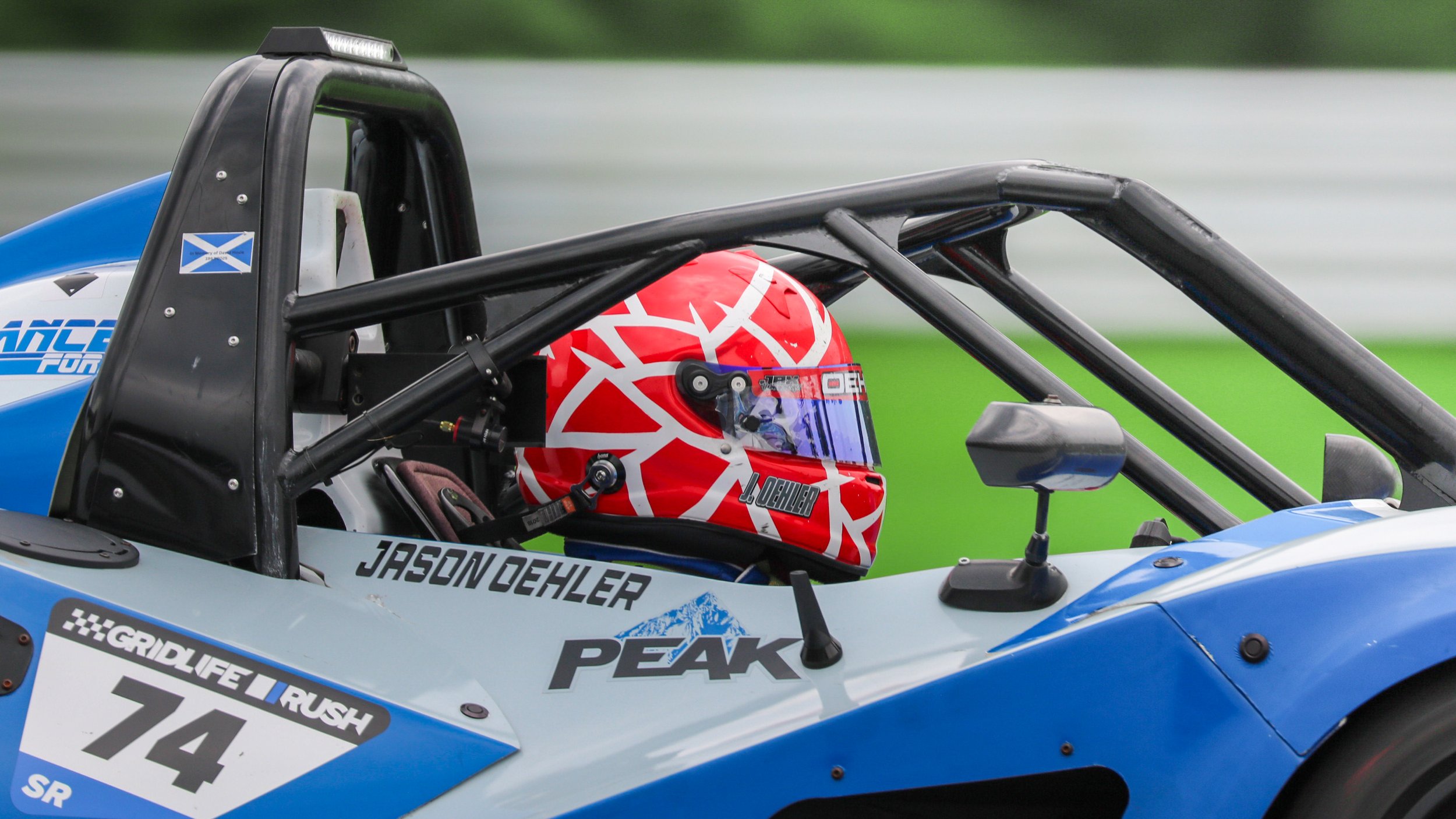 Close-up of a race car driver inside a blue open-wheel race car, wearing a red and white geometric helmet, with various sponsor stickers and the name Jason Dehler on the body of the car.