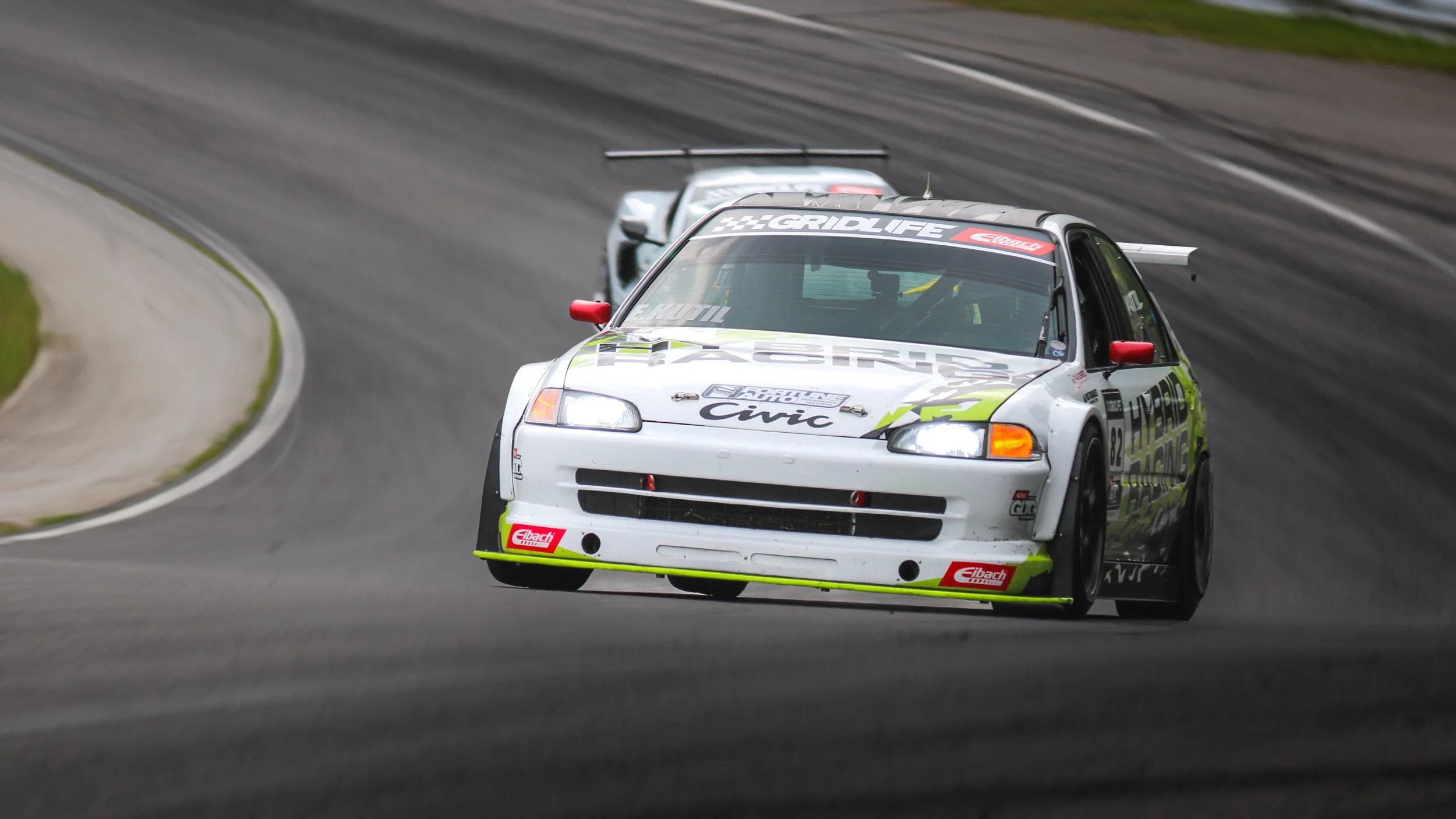 Race cars on a racing track, with the car in front being a white and gray Honda Civic rally car leading, followed by a silver car, on a curved section of the track.