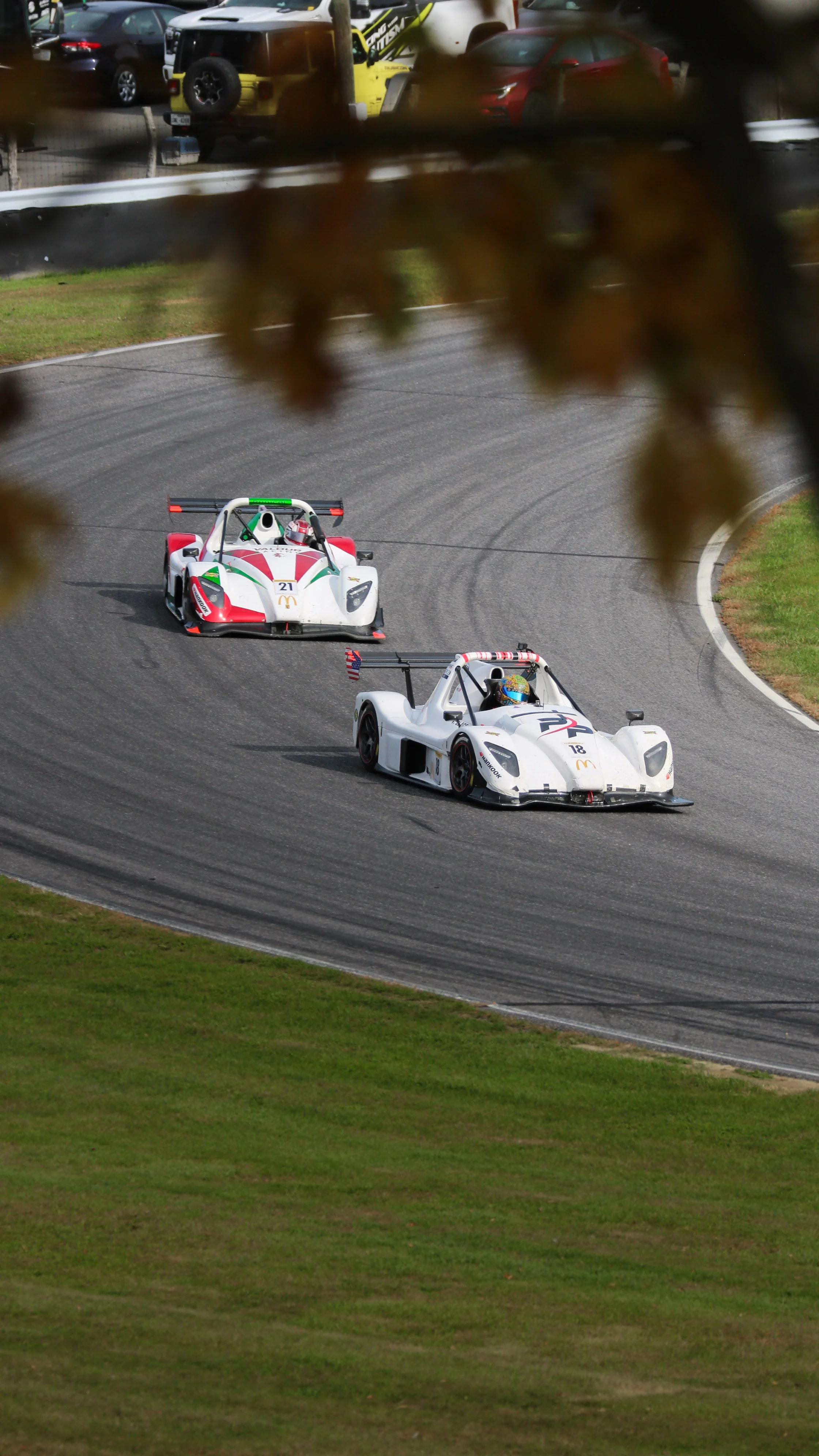 Two race cars speeding around a curved race track, with a background of parked cars and autumn leaves partially covering the view.