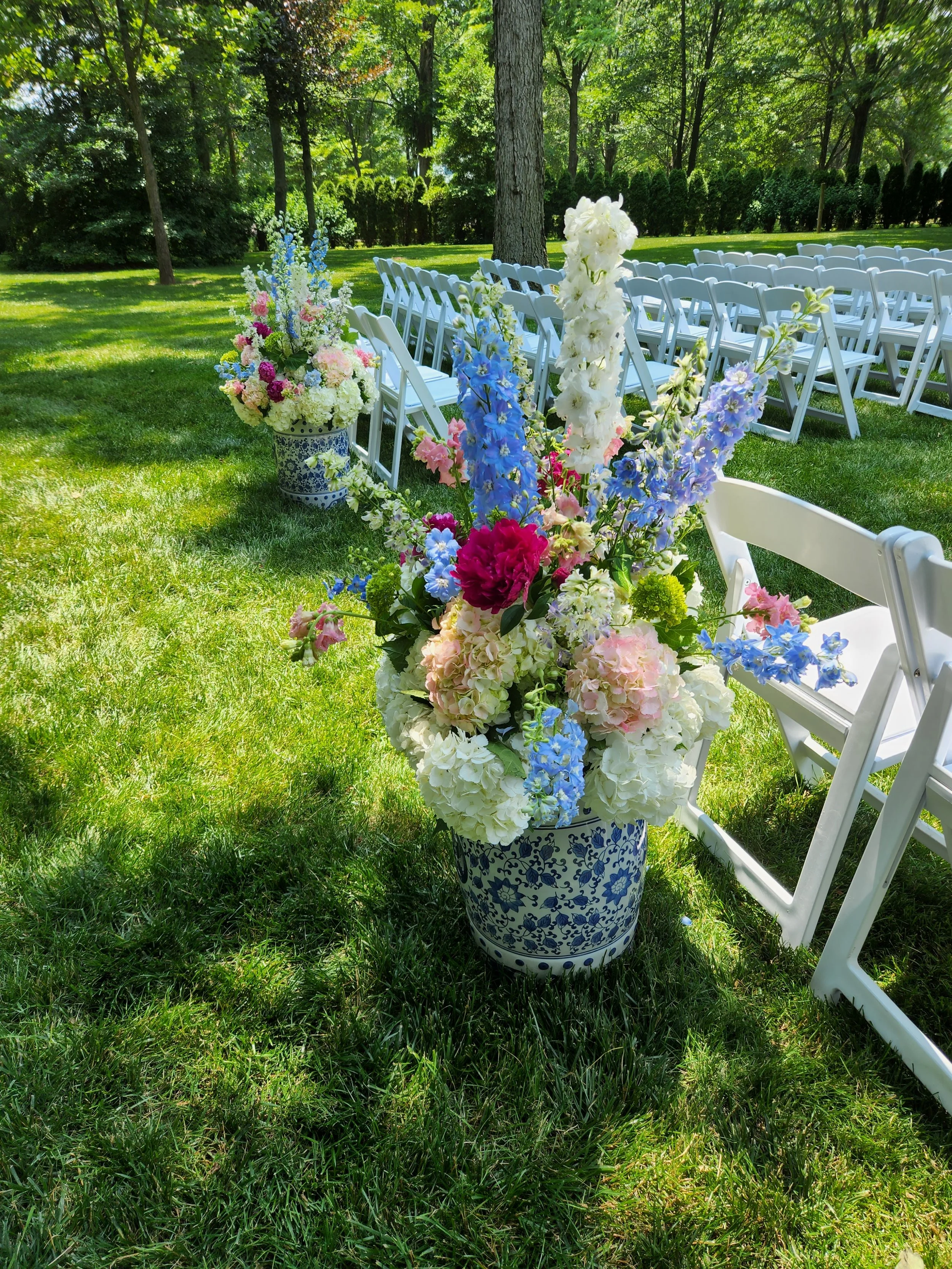 Aisle arrangements with white and pink hydrangeas, magenta peonies, and blue delphinium atop blue and white Oriental porcelain garden seats for a wedding ceremony.