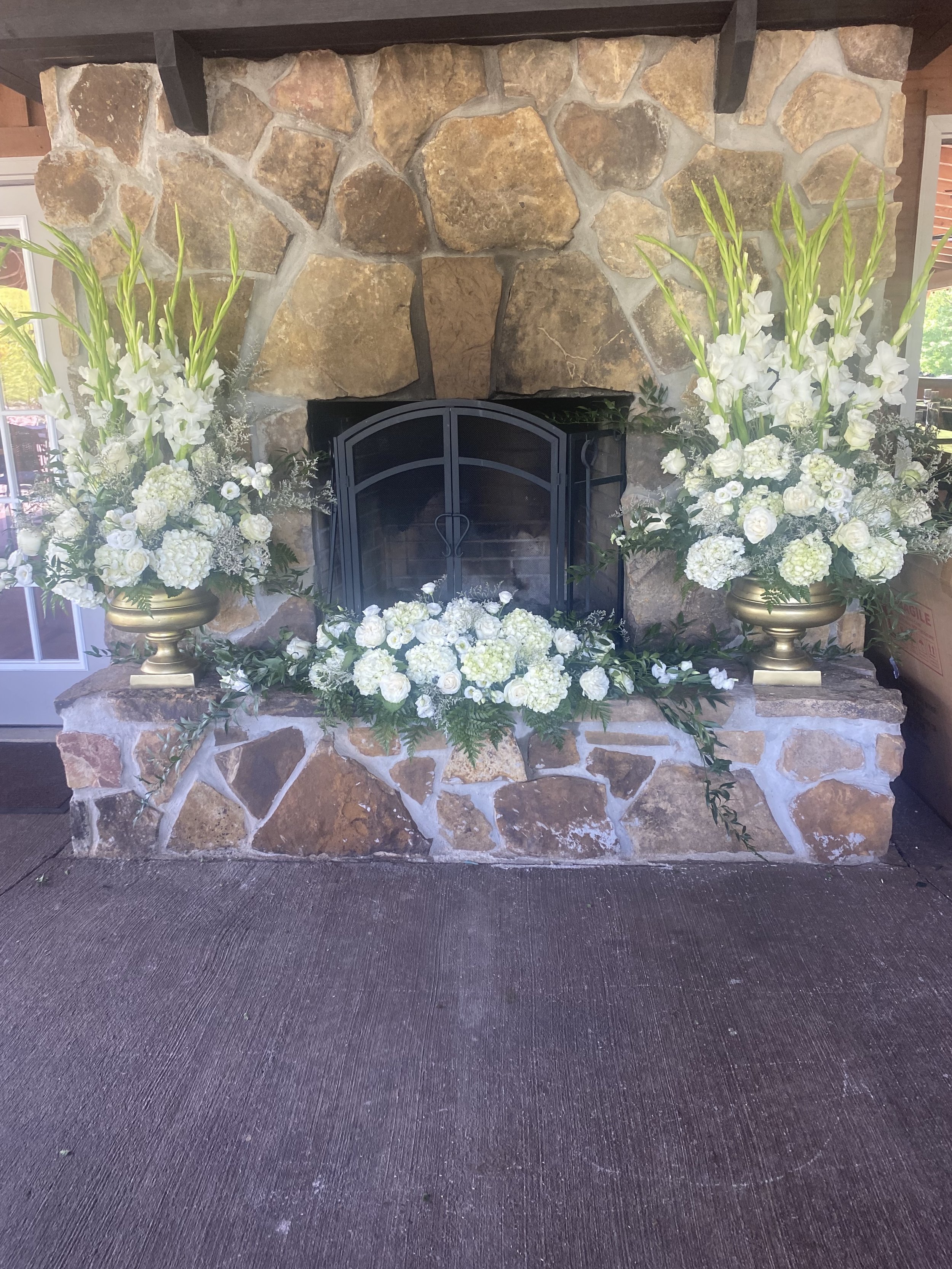 White floral arrangements with tall gladiolus, hydrangeas, roses, and lisianthus in gold urns on either side of a stone fireplace, with a white flower garland across the hearth for a wedding reception.