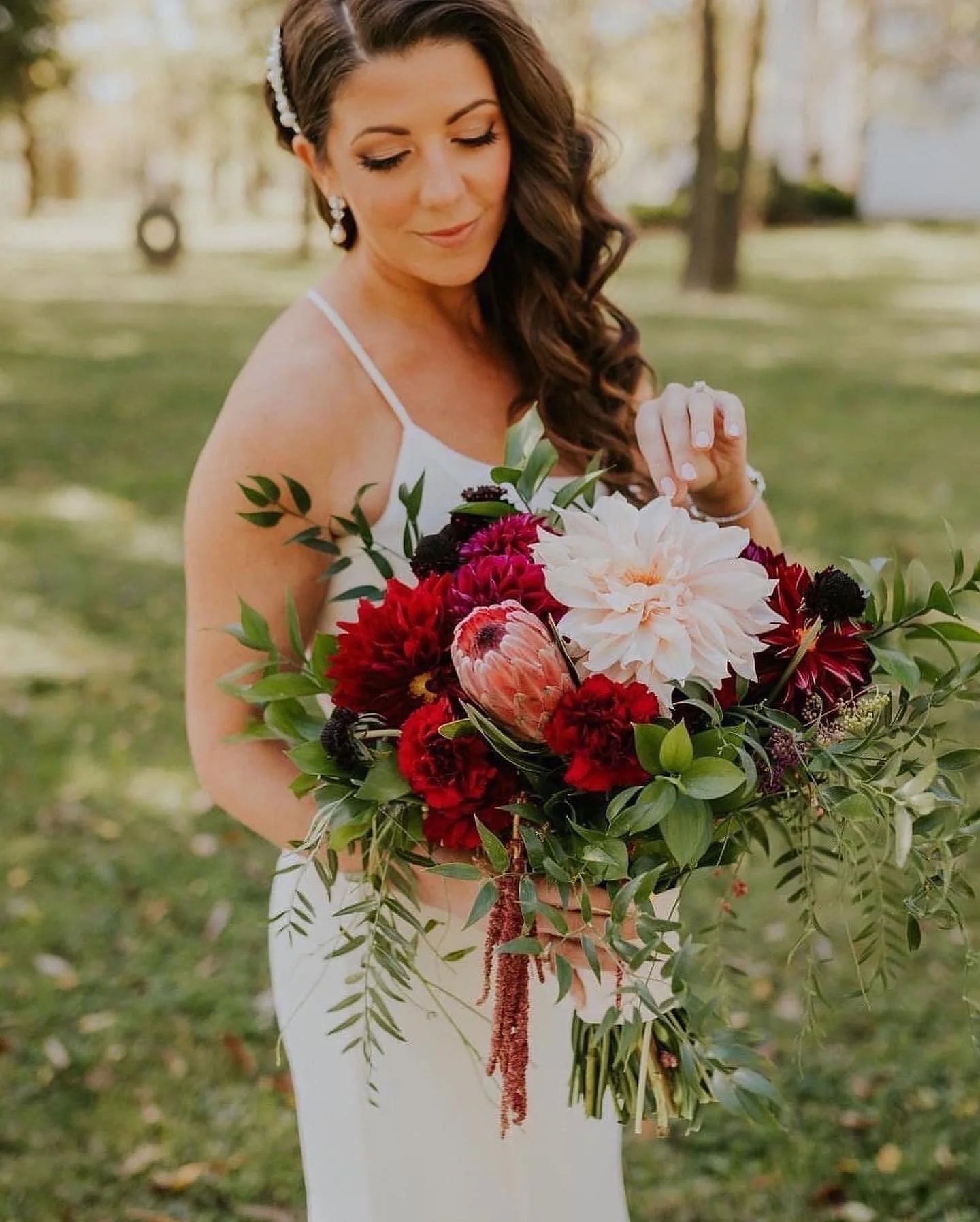 A bride with long brown hair in loose curls, wearing a white sleeveless dress and a pearl headband, holding a large bouquet of mixed red, pink, and white flowers and greenery outdoors.