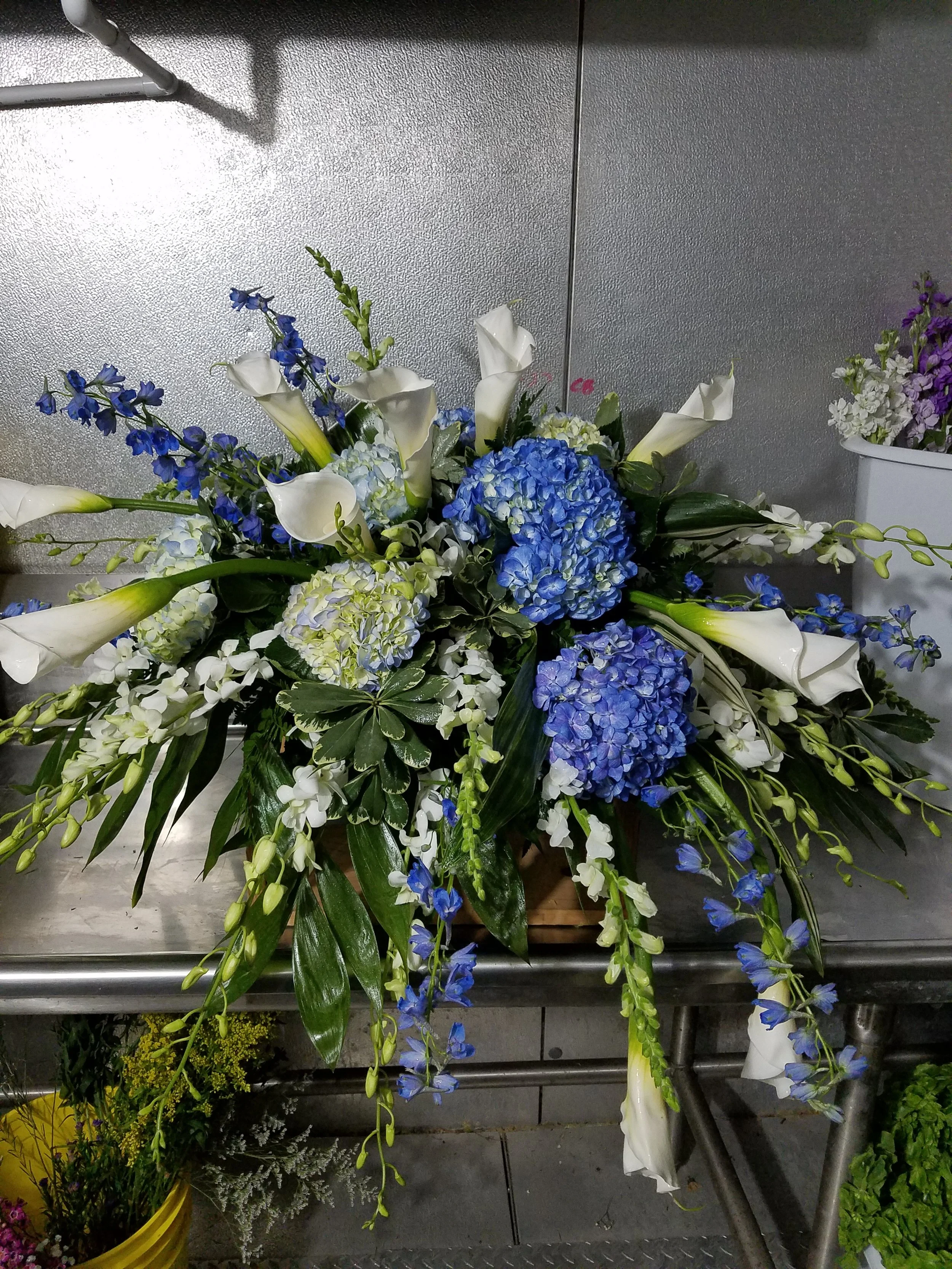 A casket spray featuring white calla lilies, blue hydrangeas, and blue delphiniums, with various green foliage.