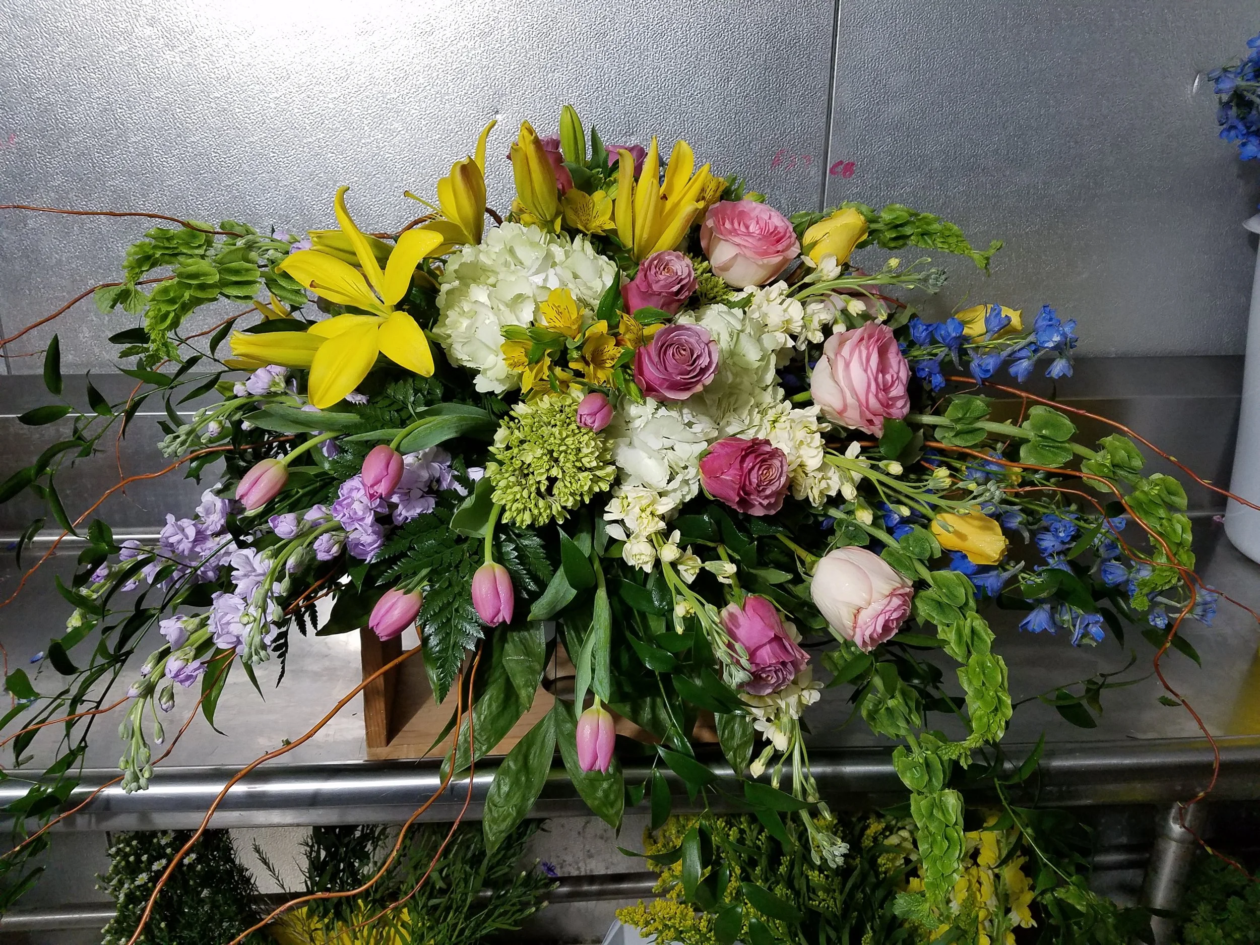 A colorful casket spray of various flowers including yellow lilies, pink roses, white hydrangeas, purple tulips, and blue delphiniums, with greenery and some branches.