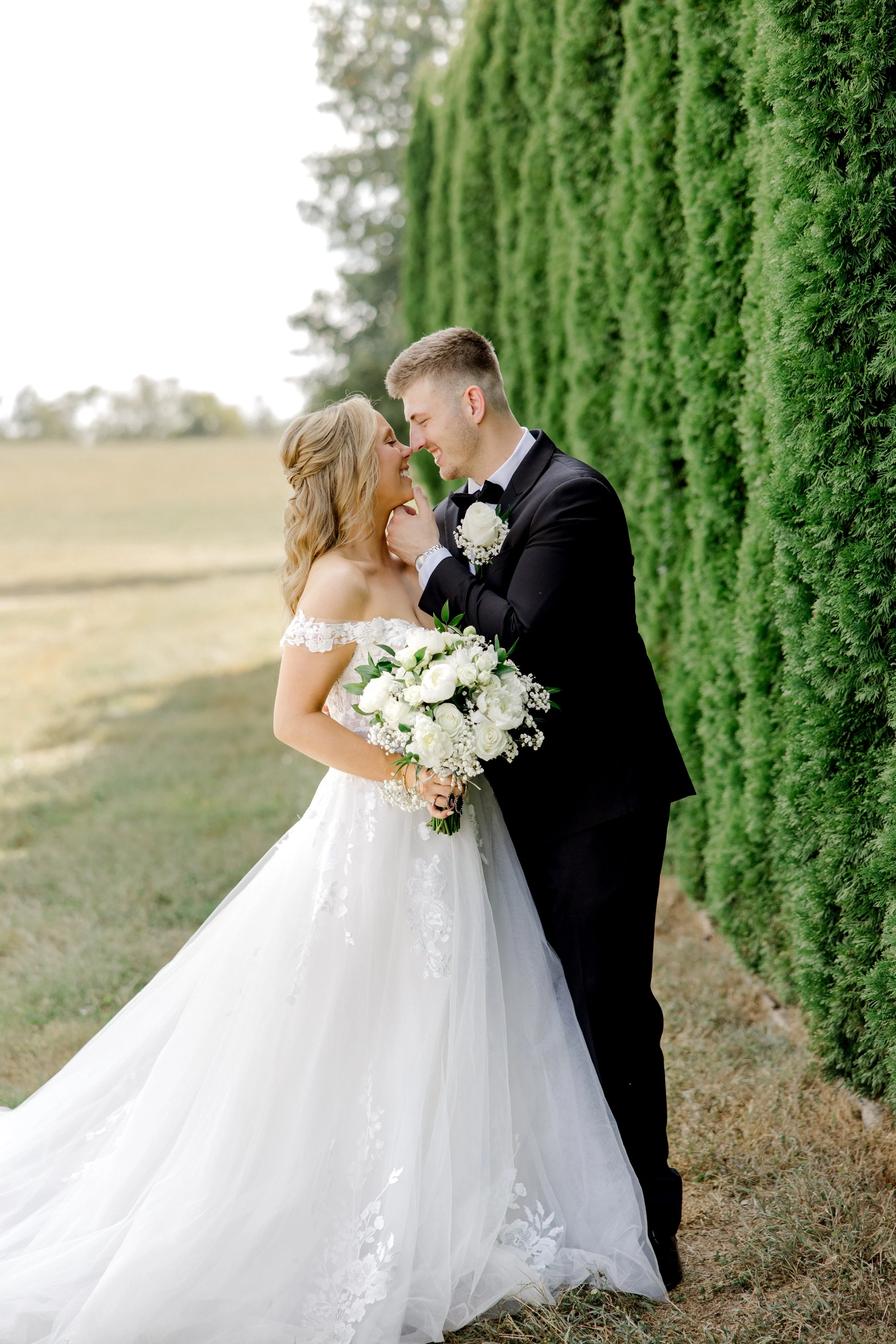 A bride carrying a hand tied bouquet of white peonies, roses, and baby's breath and groom wearing a white rose boutonniere.