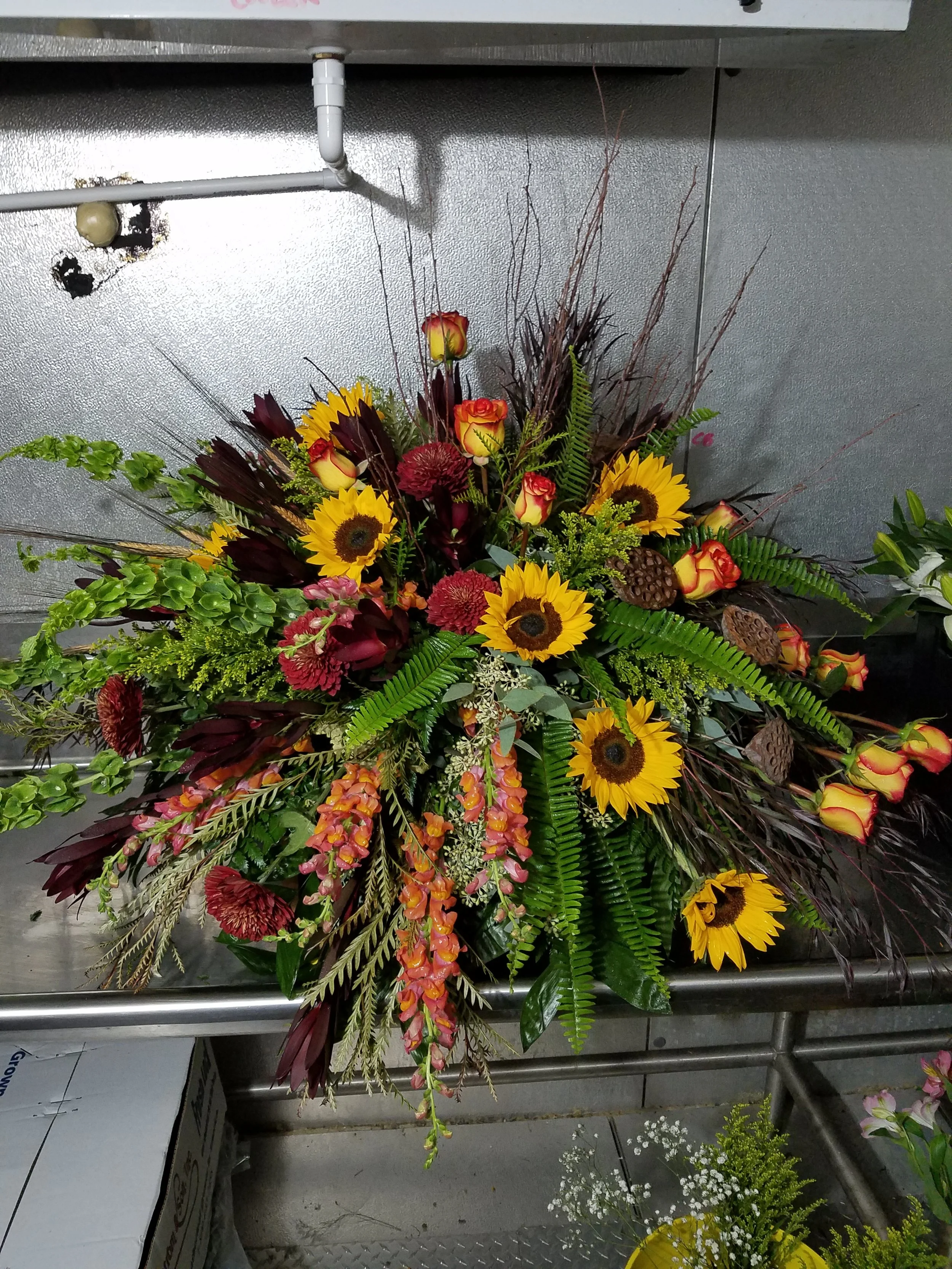 A colorful autumnal casket spray featuring sunflowers, roses, red chrysanthemums, orange snapdragons, greenery, and decorative seed pods.