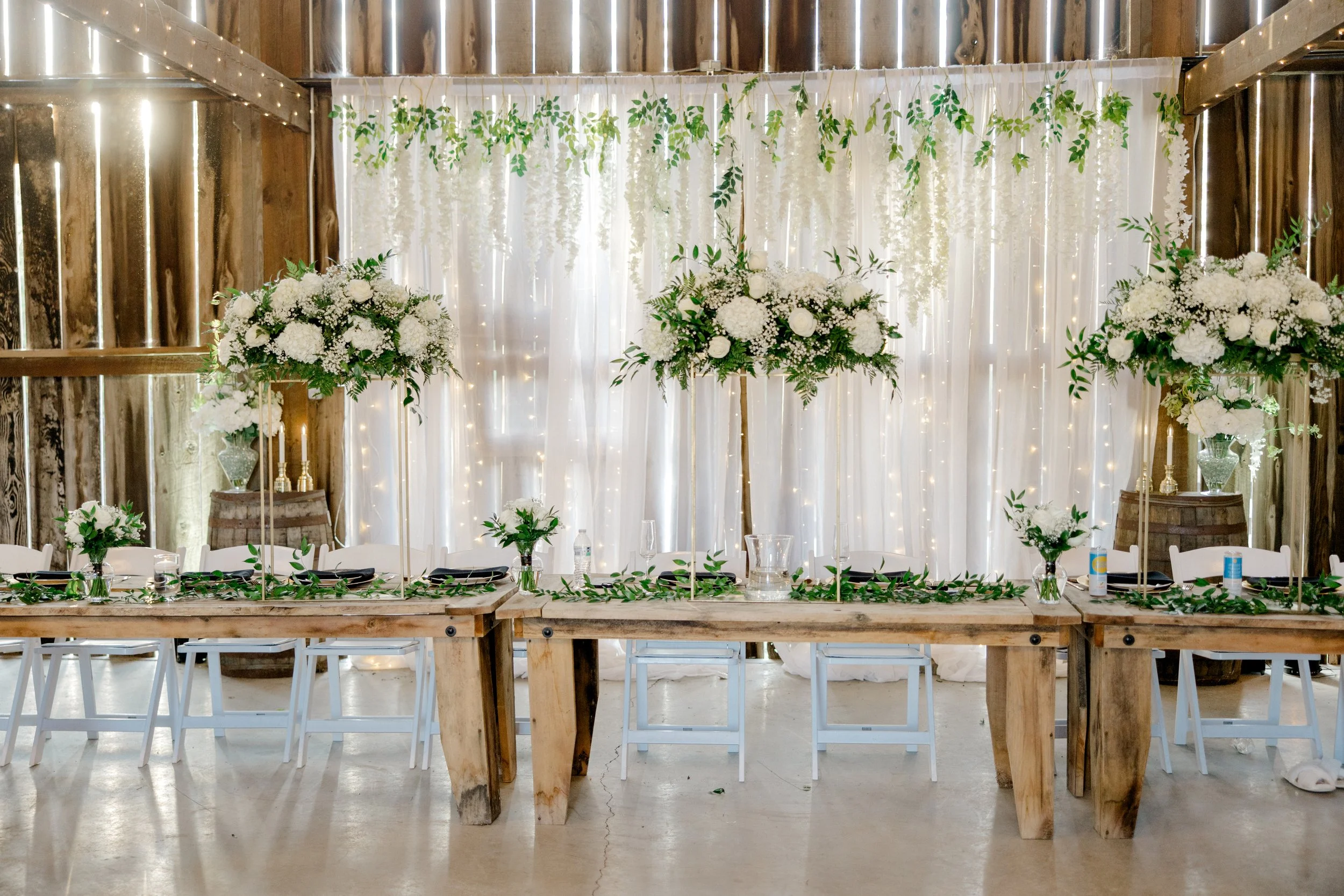 Elegant wedding reception table with white floral centerpieces and greenery, set against a backdrop of white curtains with hanging white flowers and string lights, housed in a rustic venue.