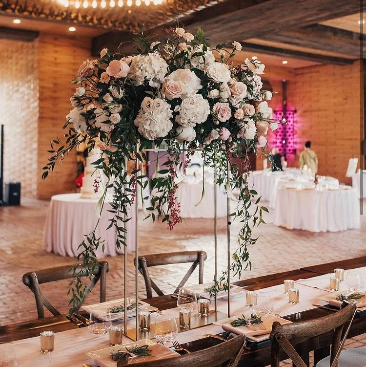 A large floral centerpiece with white and pale pink roses, white hydrangeas, and greenery on a tall stand decorates a dining table at a wedding reception.