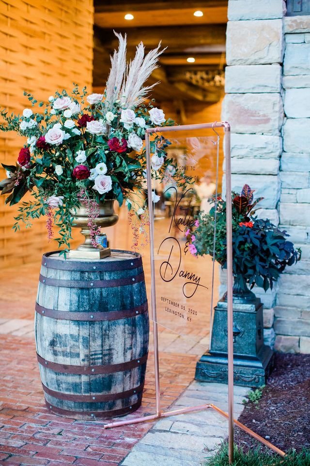 A floral arrangement with roses, lisianthus, pampas grass, and greenery on a wooden barrel adorning the entrance to a wedding ceremony.