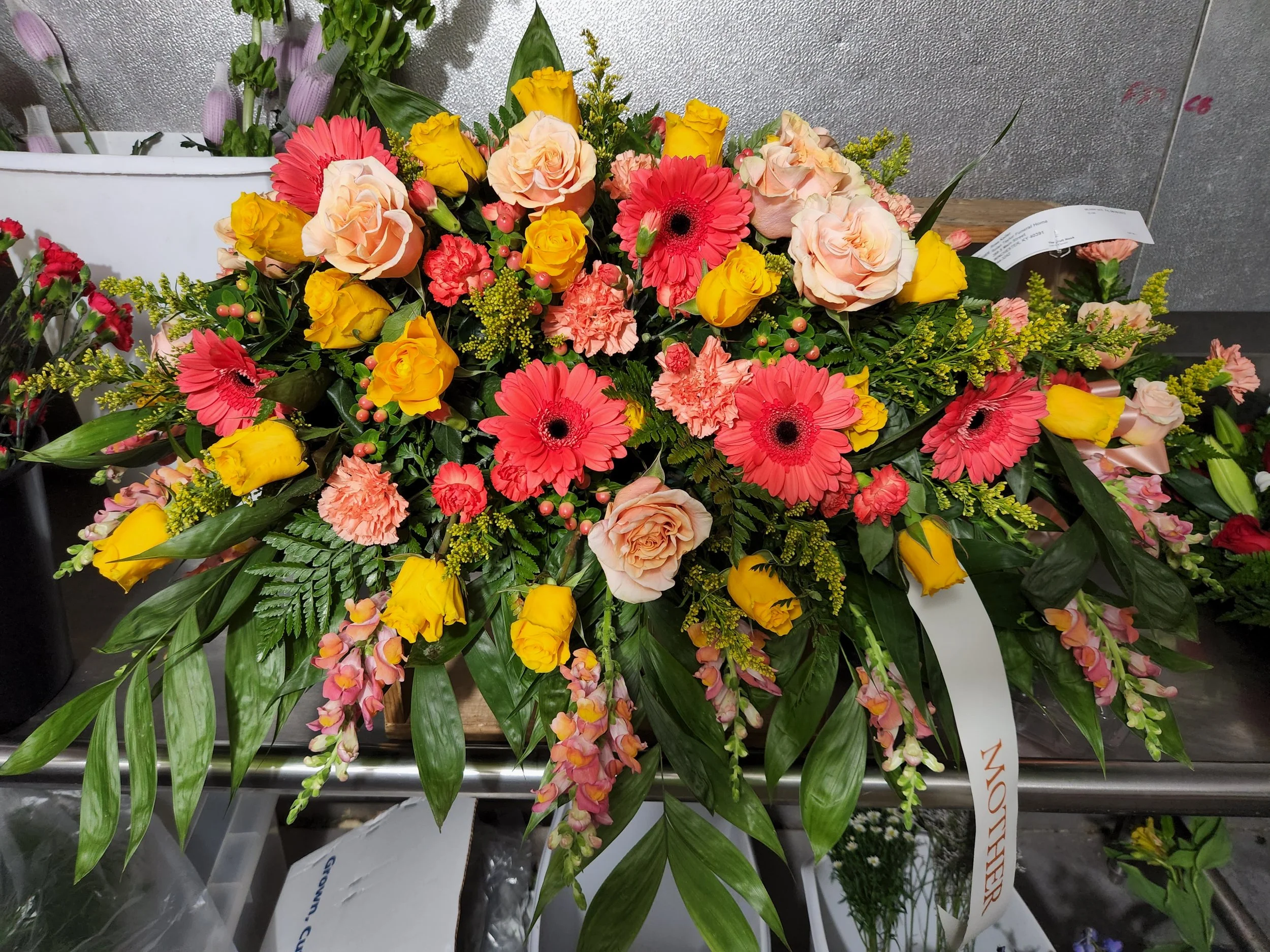 Colorful casket spray with pink gerbera daisies, yellow roses, and light pink roses, surrounded by green foliage.