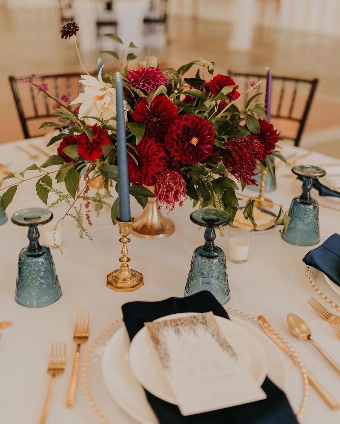 Elegant table centerpiece with red, pink, and white dahlias in a gold vase, surrounded by blue candles.