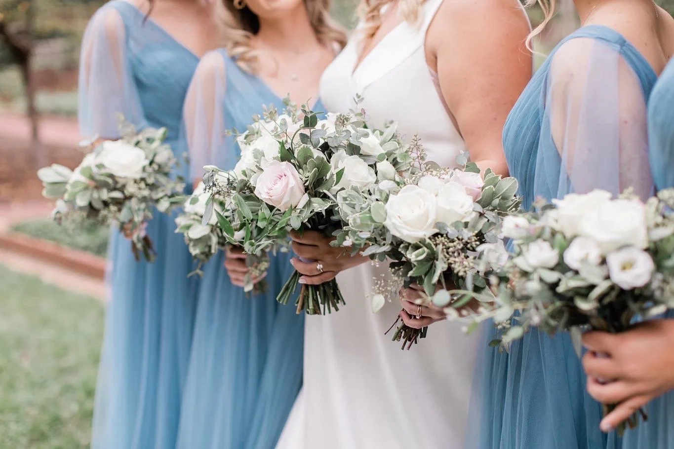 Close-up of a bride and bridesmaids holding bouquets of white and pink roses and greenery during an outdoor wedding.