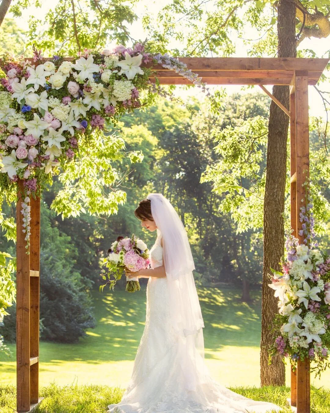 Bride in a white wedding gown and veil holding a bouquet of pink, white, and purple flowers, standing under a wooden arch decorated with flowers and greenery during a sunny outdoor wedding ceremony.