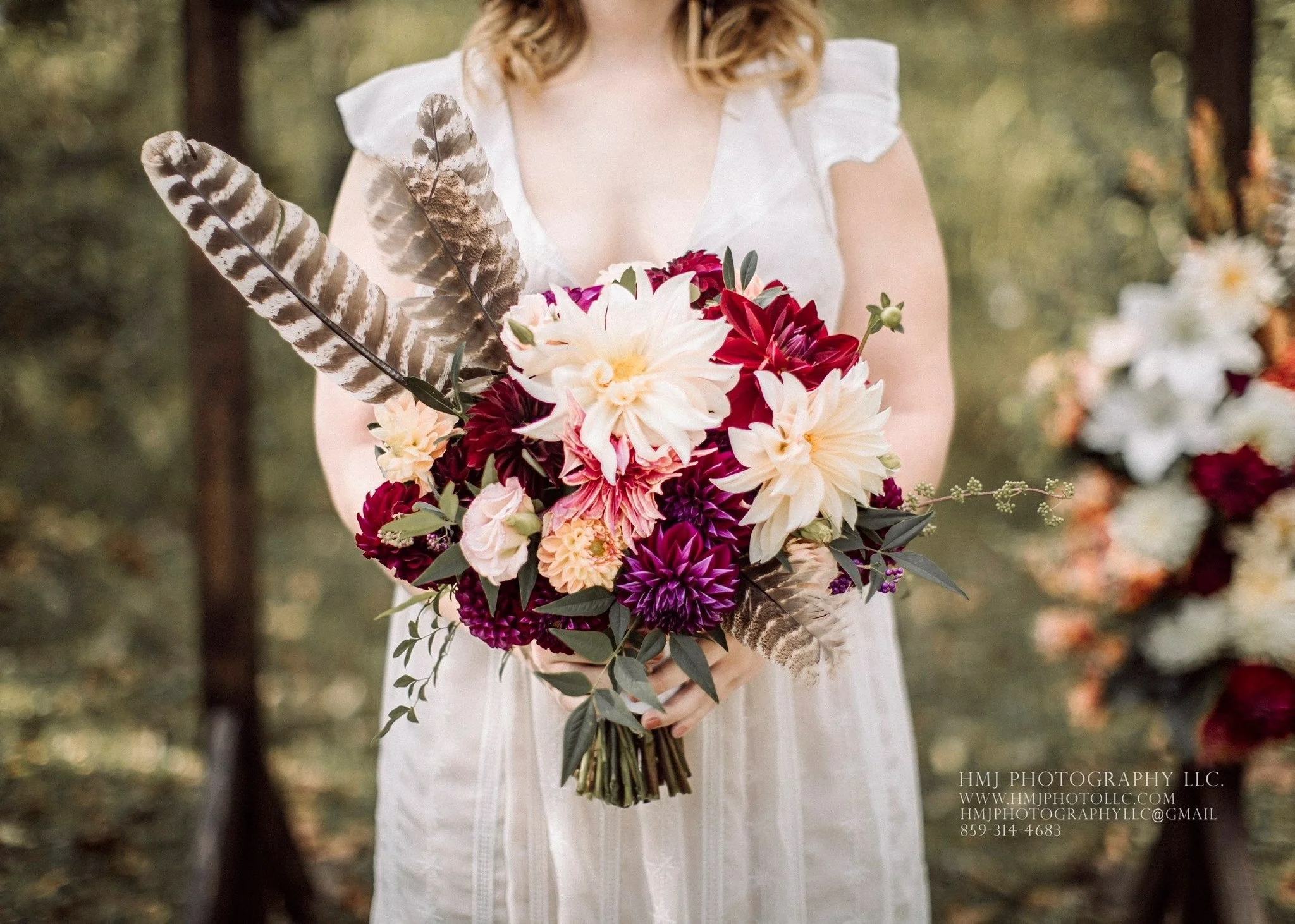 A colorful bouquet of flowers, including large white, maroon, purple, and peach dahlias and lisianthus, accented with green leaves and feathers.