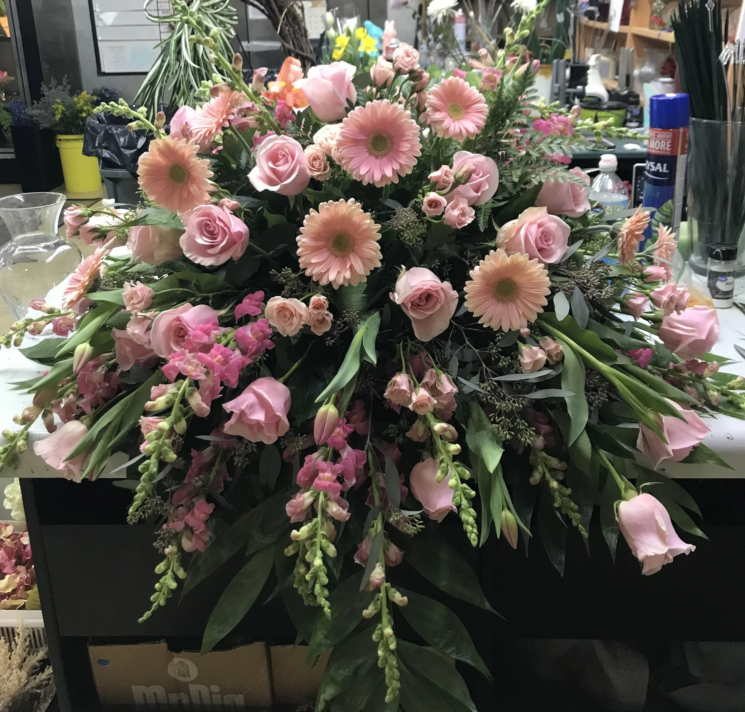 A casket spray of pink roses, gerbera daisies, and snapdragons with green foliage.