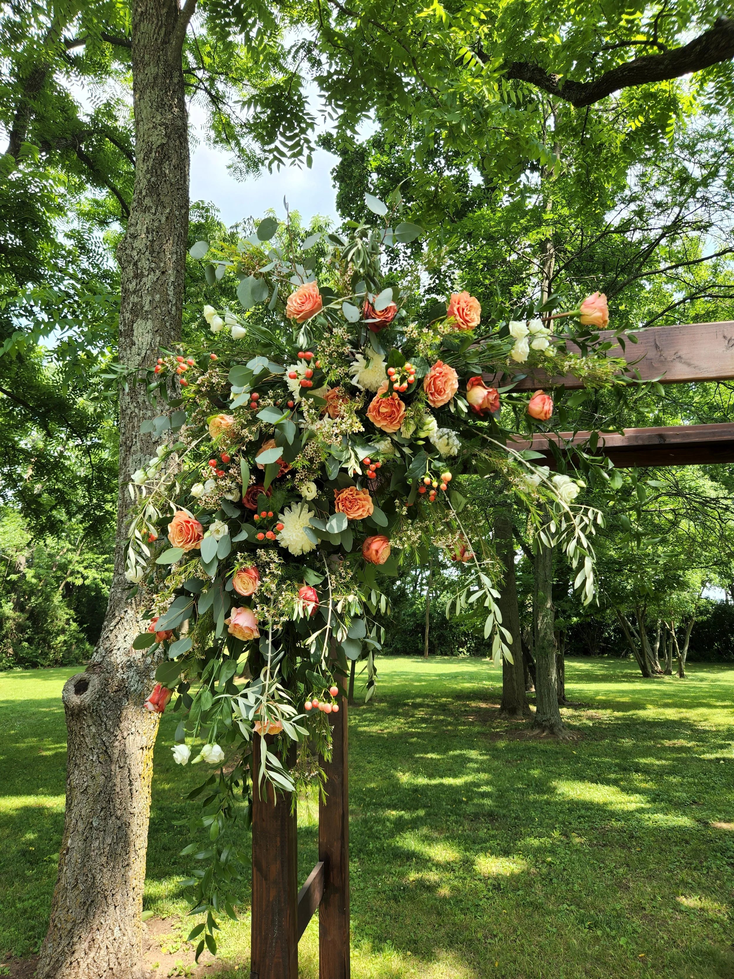 A floral swag with peach, orange and white roses, green leaves, and hypericum berries, decorating a wooden arbor for a wedding ceremony.