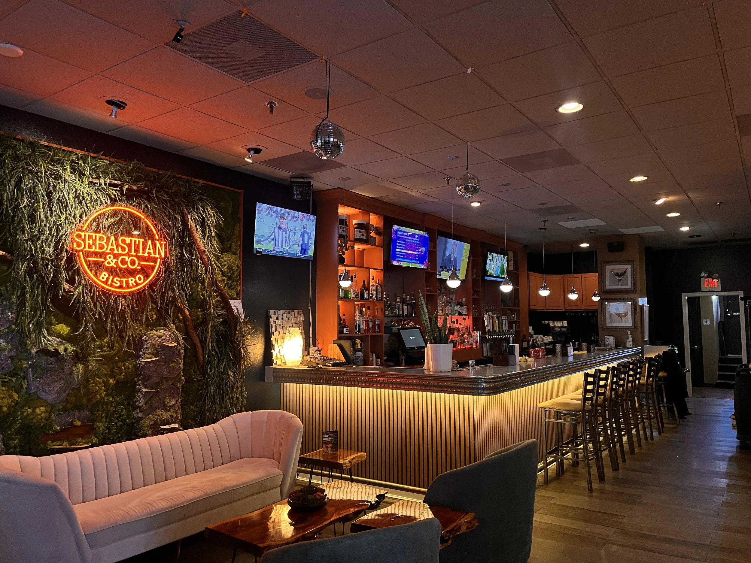 A view of a cozy bar and restaurant area with a wooden bar counter, multiple televisions displaying sports, and bar stools. There is a neon sign that reads 'SEBASTIAN & CO. BISTRO' on a wall decorated with greenery. The seating includes a pink velvet couch and various chairs, while disco balls hang from the ceiling.